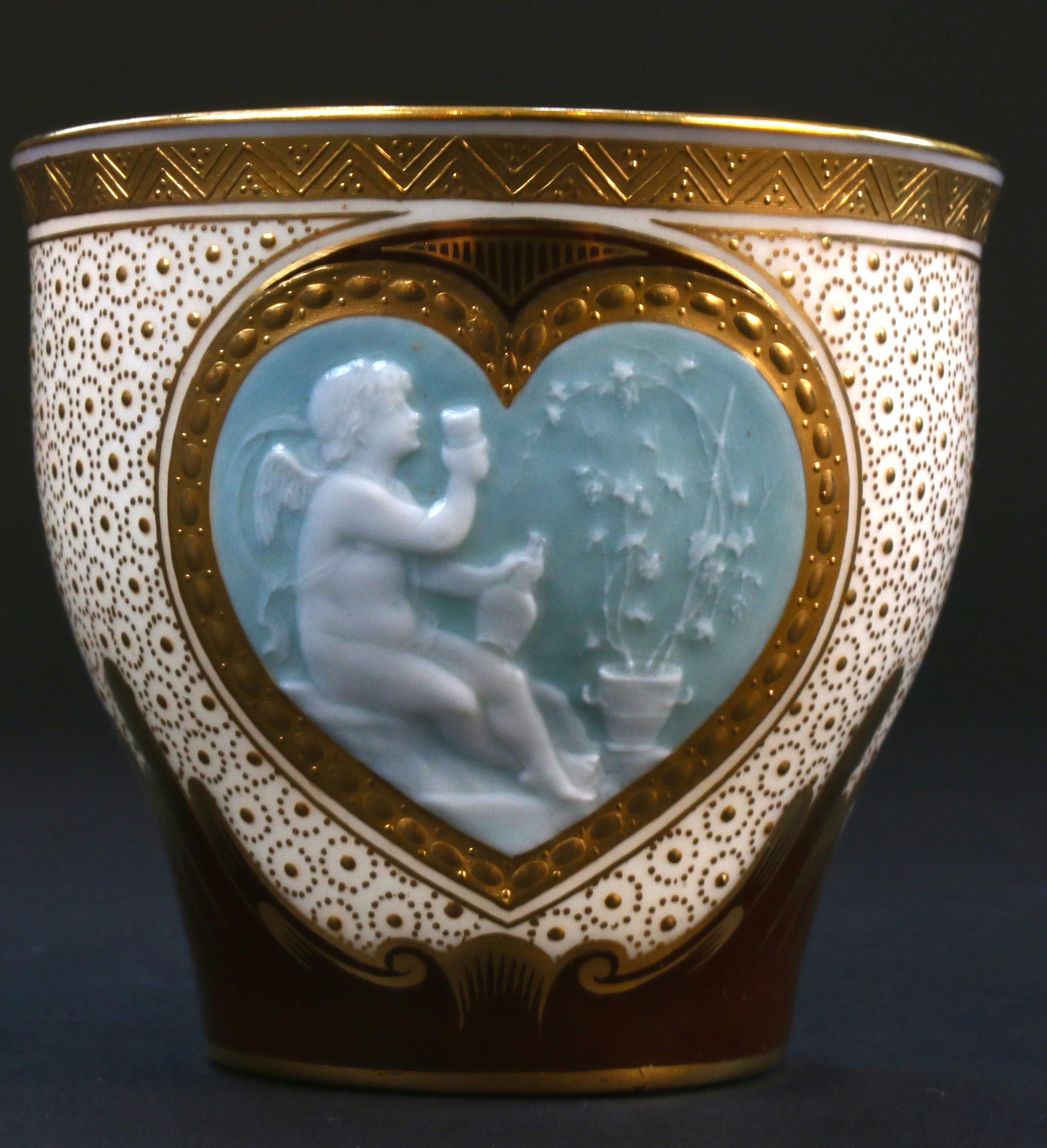 This rarely available Minton pate sur pate cup would make a wonderful display item or addition to a collection. The cup contains a 22-karat gold outlined heart-shaped pate sur pate plaque, rendered with white slip on a blue background, featuring a