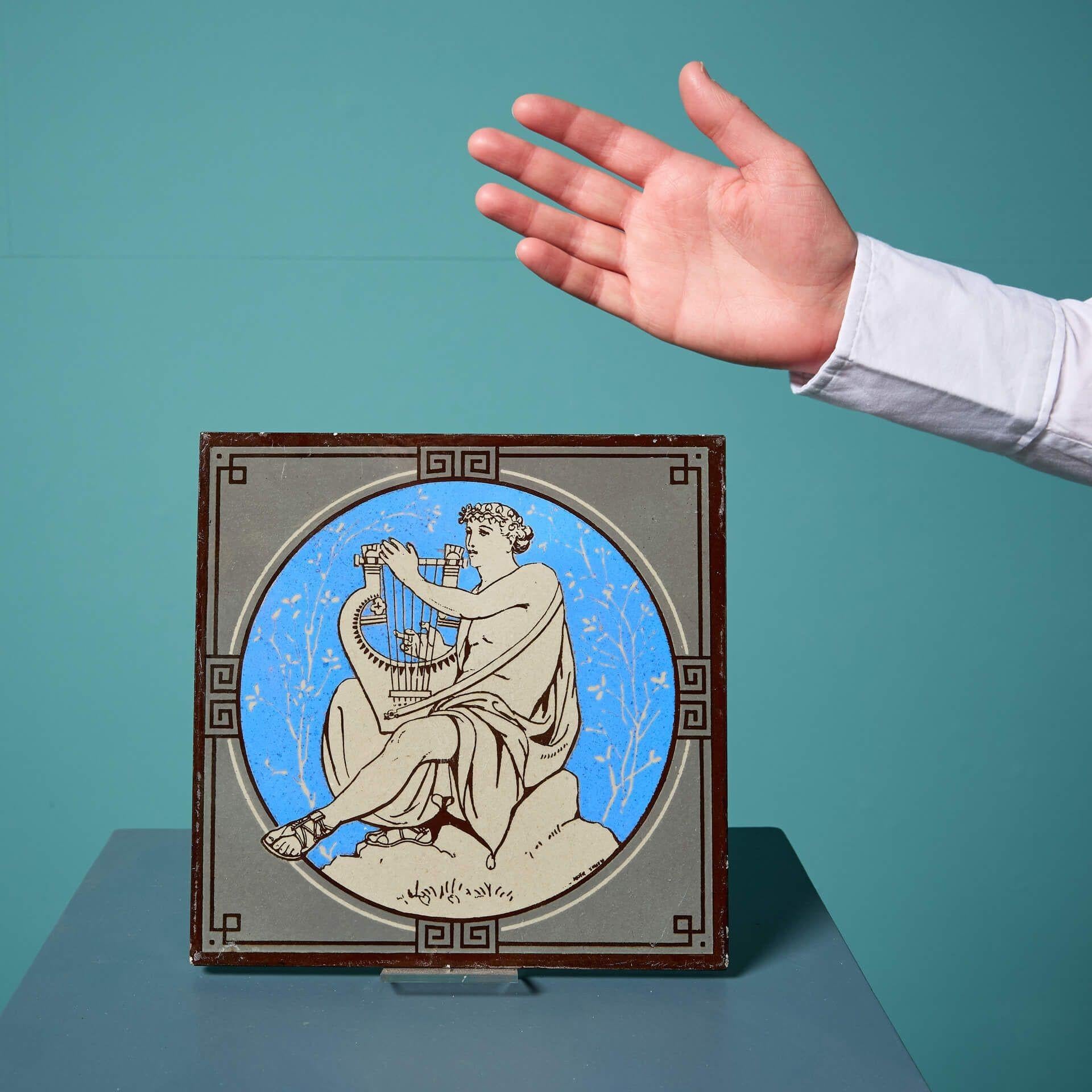 An antique Minton tile from the series ‘Classical Figures with Musical Instruments’ designed by the artist John Moyr Smith.

This 8 inch tile depicts a woman in noble dress playing a harp on a bright blue background surrounded by a stylised