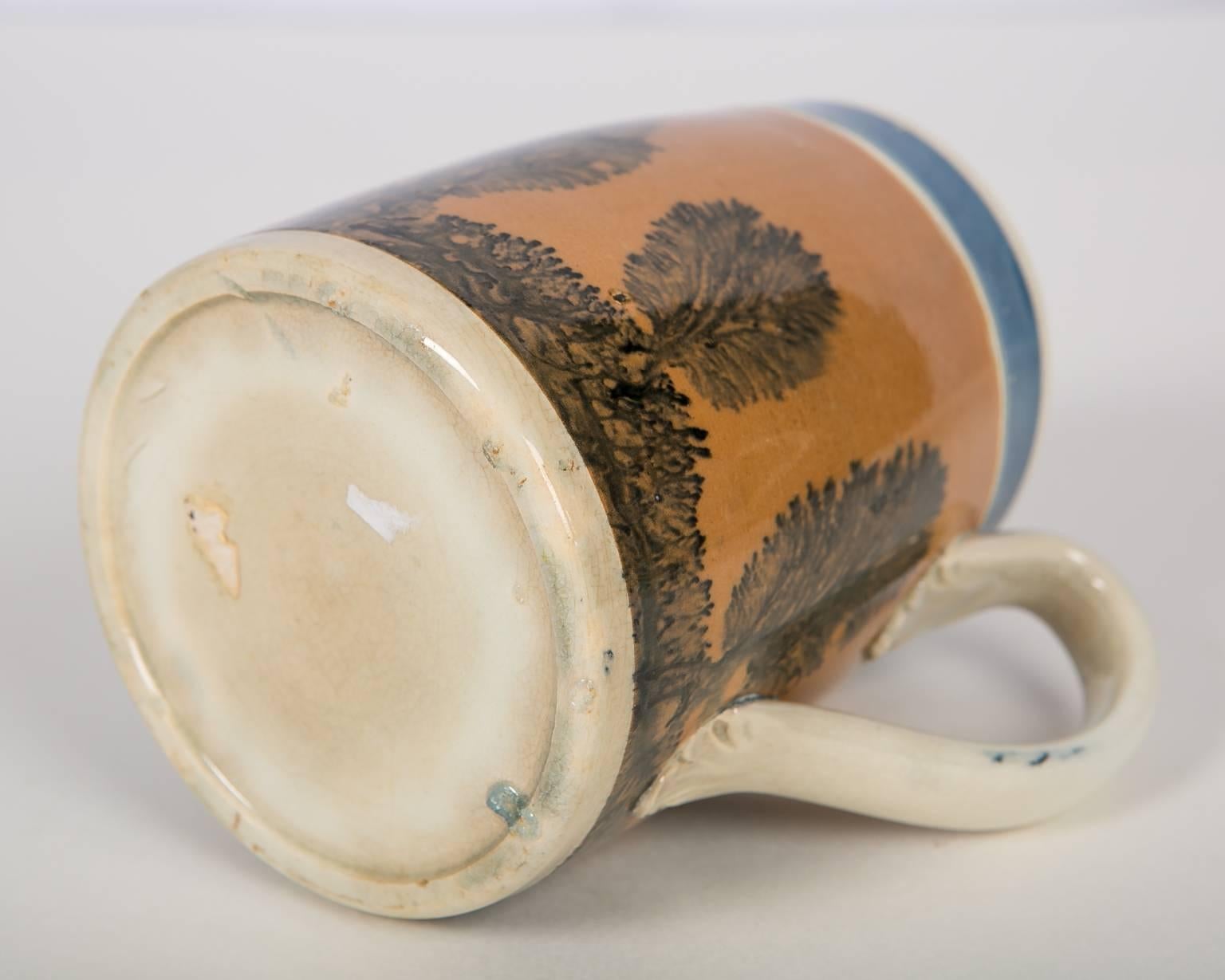 A Mochaware half pint mug molded in an elegant shape with a wide band of burnt orange slip decorated with vertical tree-like dendritic designs. The 