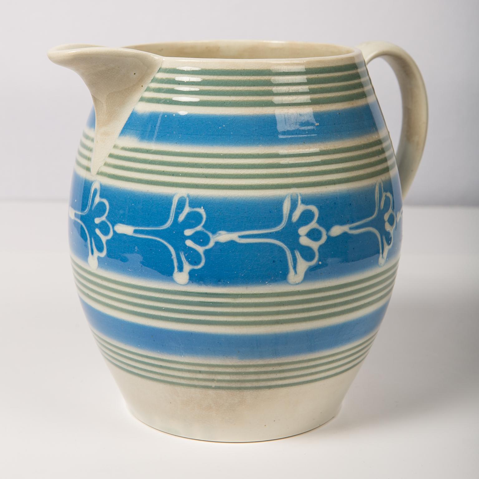 We are pleased to offer this large antique mochaware pitcher made in England circa 1830. It is colored with bands of powder blue and olive green slip with an offset decoration of a flower in white slip. In 