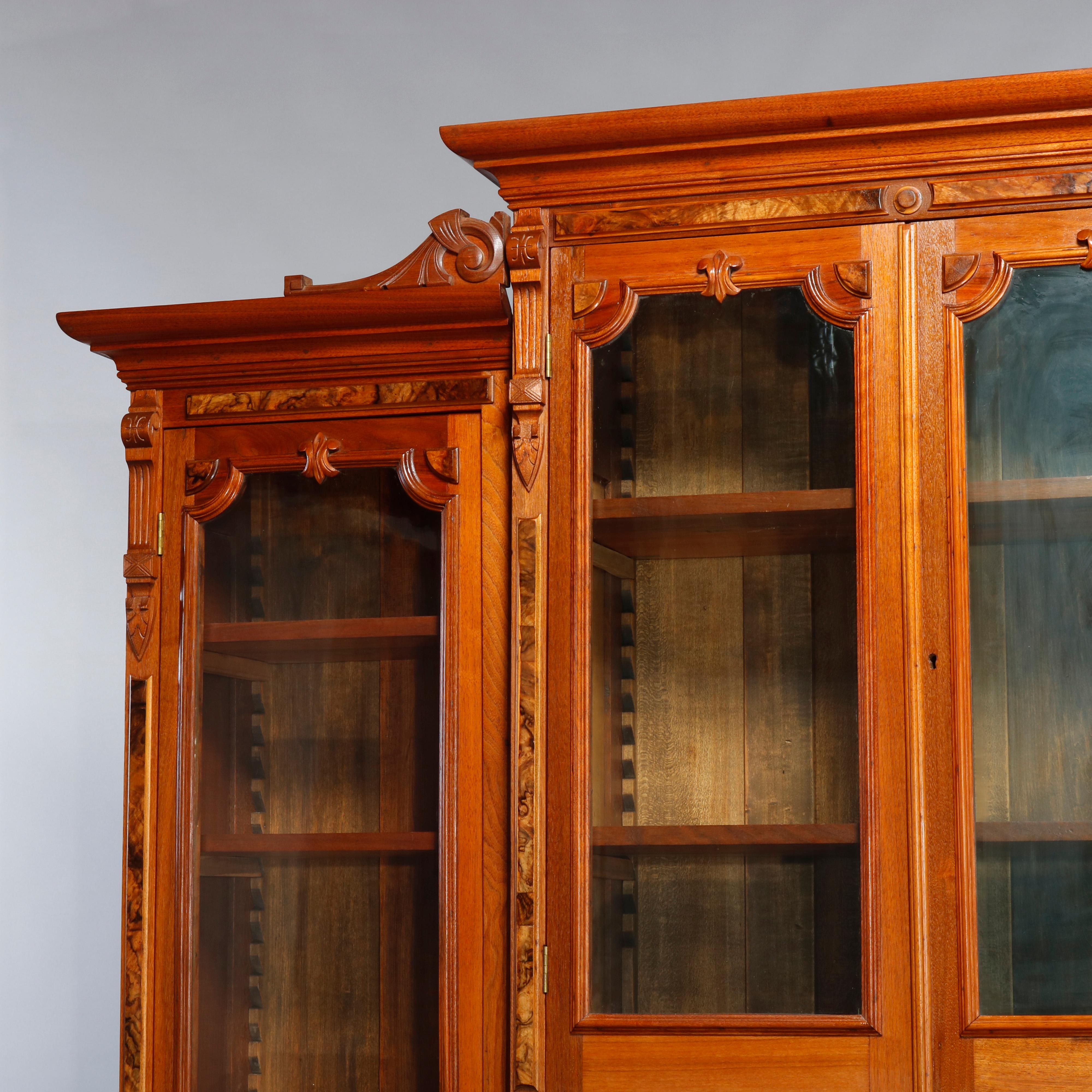 An antique and monumental Eastlake step-back secretary bookcase offers carved walnut and burl construction with central case having upper bookcase with double glass doors over drop front desk and drawer tower, flanked by set back tall bookcases each