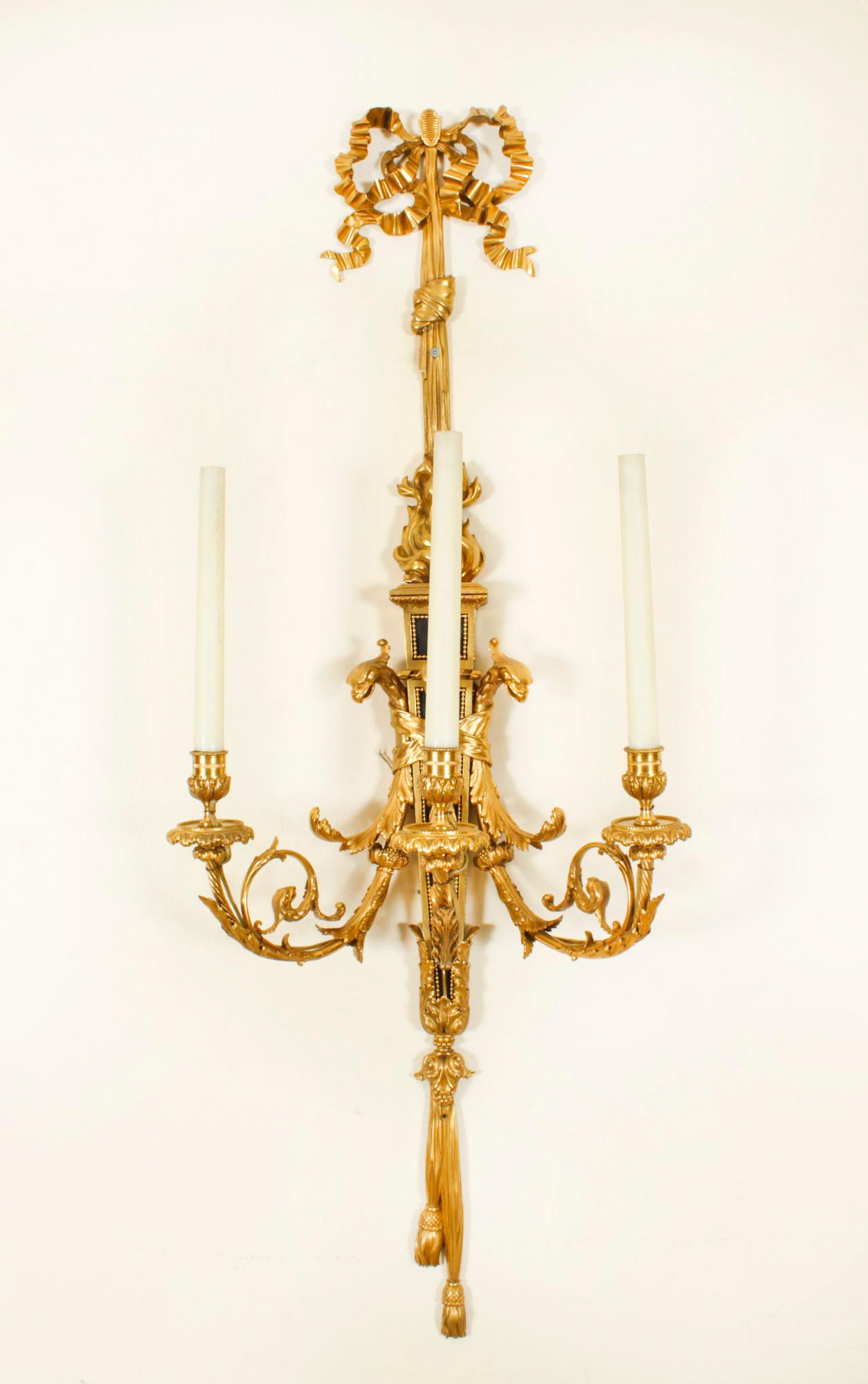 This is a monumental pair of antique French Louis XVI Revival gilt and patinated bronze three branch wall appliques, late 19th century in date.

They each feature backplates surmounted with a ribbon tied bow and tassels above scrolling foliate