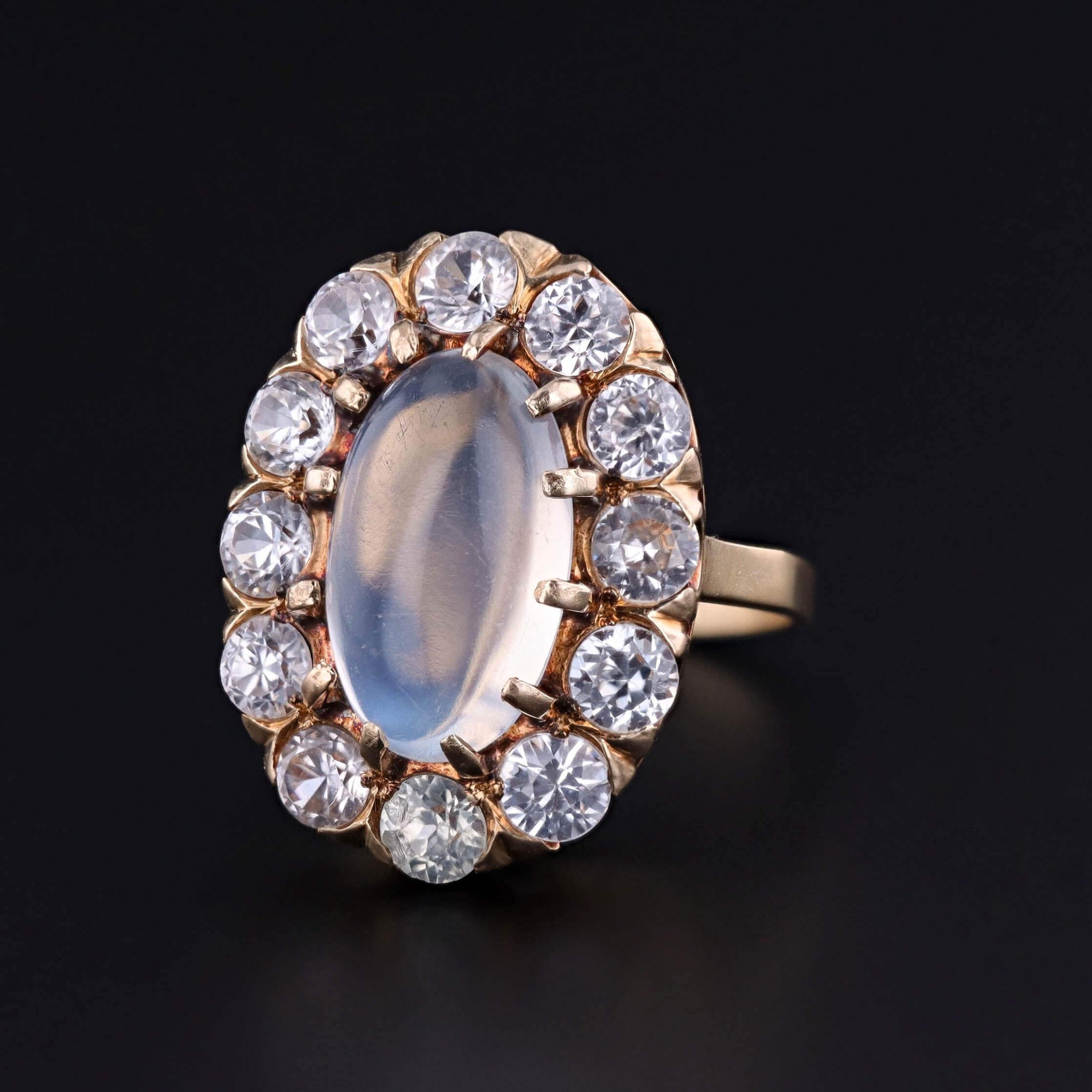 Bijou antique lumineux réimaginé pour le collectionneur moderne, cette bague est centrée sur un cabochon de pierre de lune brillant entouré d'un halo de zircons naturels, le tout serti dans un riche or 14k. La combinaison de l'éclat éthéré de la