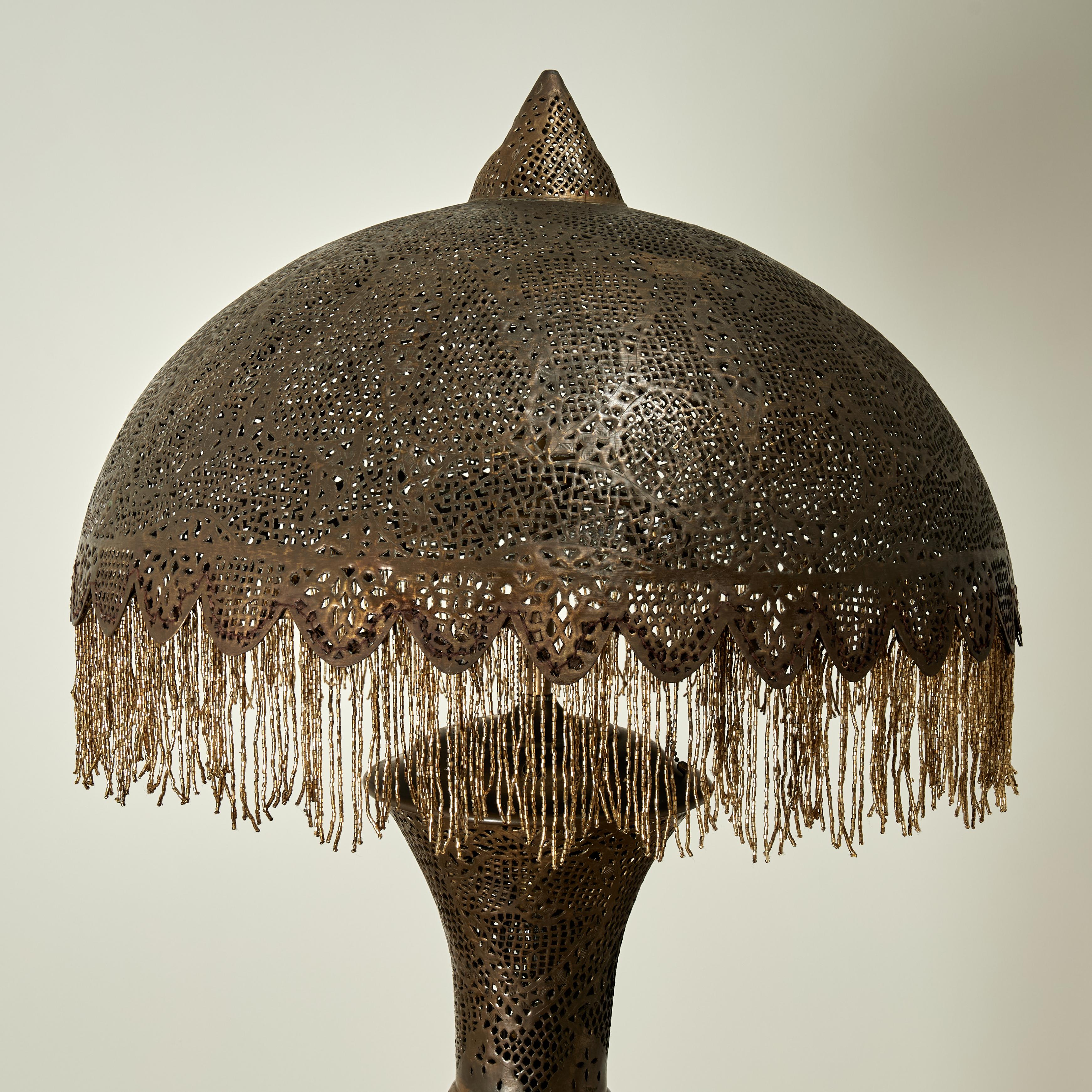 Charmant lampadaire marocain ancien en laiton perforé de motifs symboliques et floraux avec un abat-jour bombé à glands sur une colonne mauresque qui s'allume à l'intérieur sur une base ronde à trois niveaux.