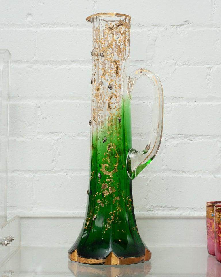 Este hermoso y ornamentado aguamanil Moser antiguo es un asombroso ejemplo de artesanía, elaborado en cristal verde a transparente con intrincados detalles dorados. El intenso color verde de la base se desvanece gradualmente hasta convertirse en