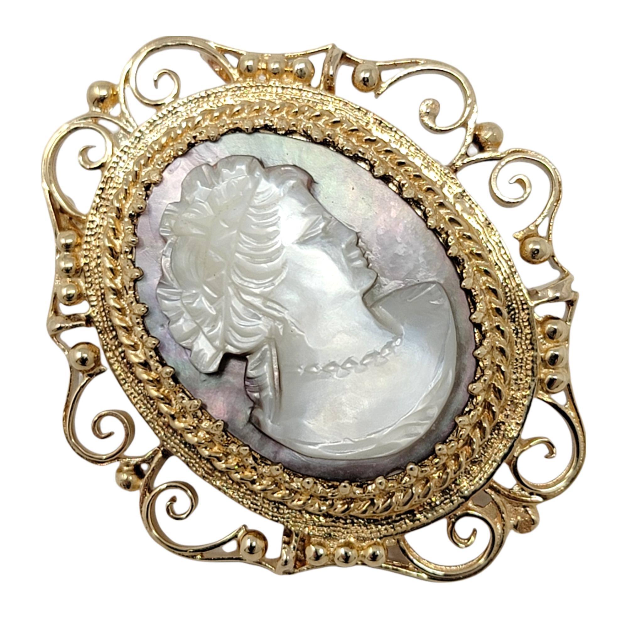 Retrocede en el tiempo con este increíble broche / colgante camafeo antiguo. Esta pieza femenina vintage presenta una Madreperla iridiscente semiopaca intrincadamente tallada, rodeada por un marco dorado texturizado. La imagen ovalada muestra el