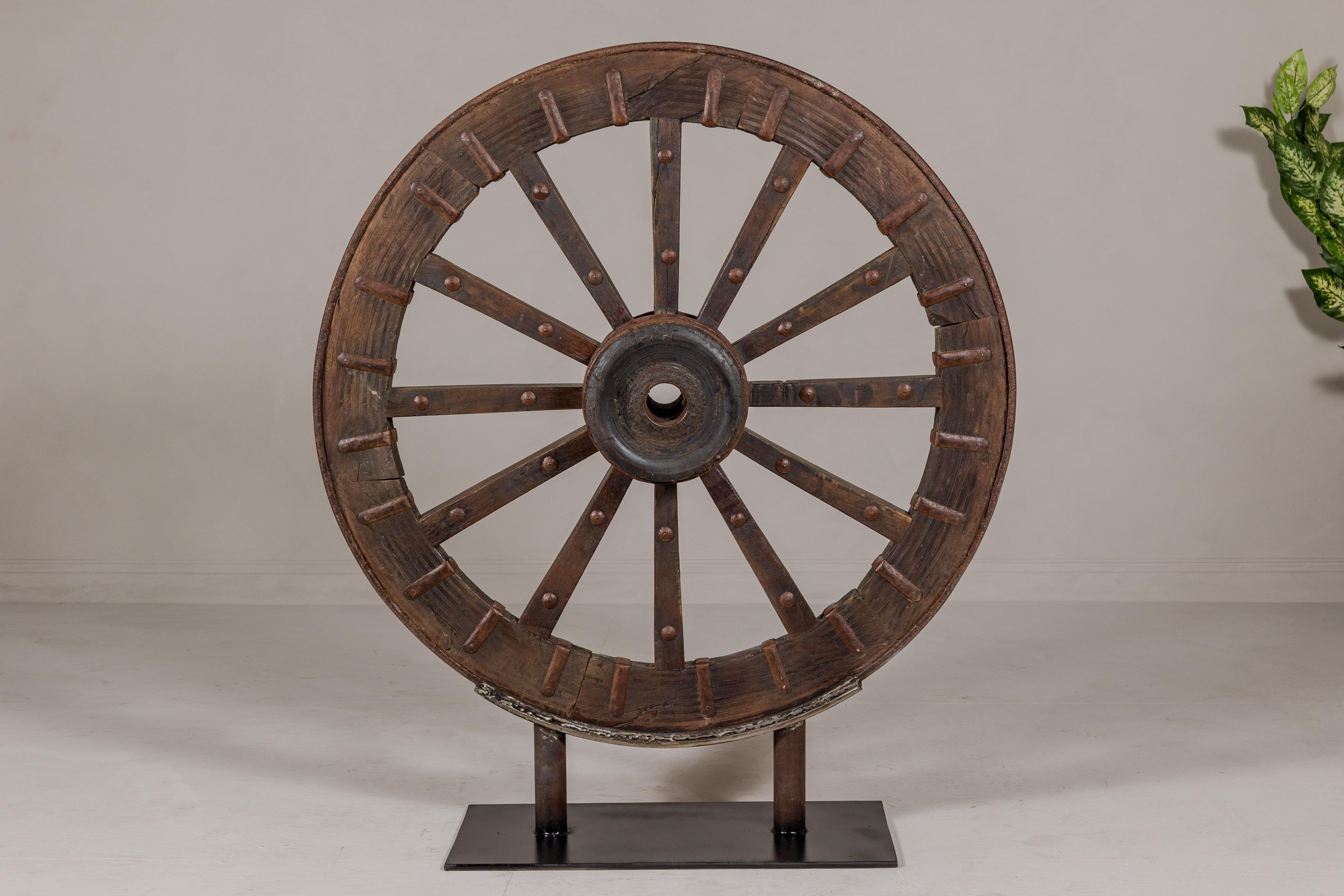 An antique Indian wood and metal cart wheel mounted on custom black metal base. Immerse yourself in the rich history and rustic charm of this antique Indian cart wheel, a unique and captivating piece that brings a touch of authenticity and timeless