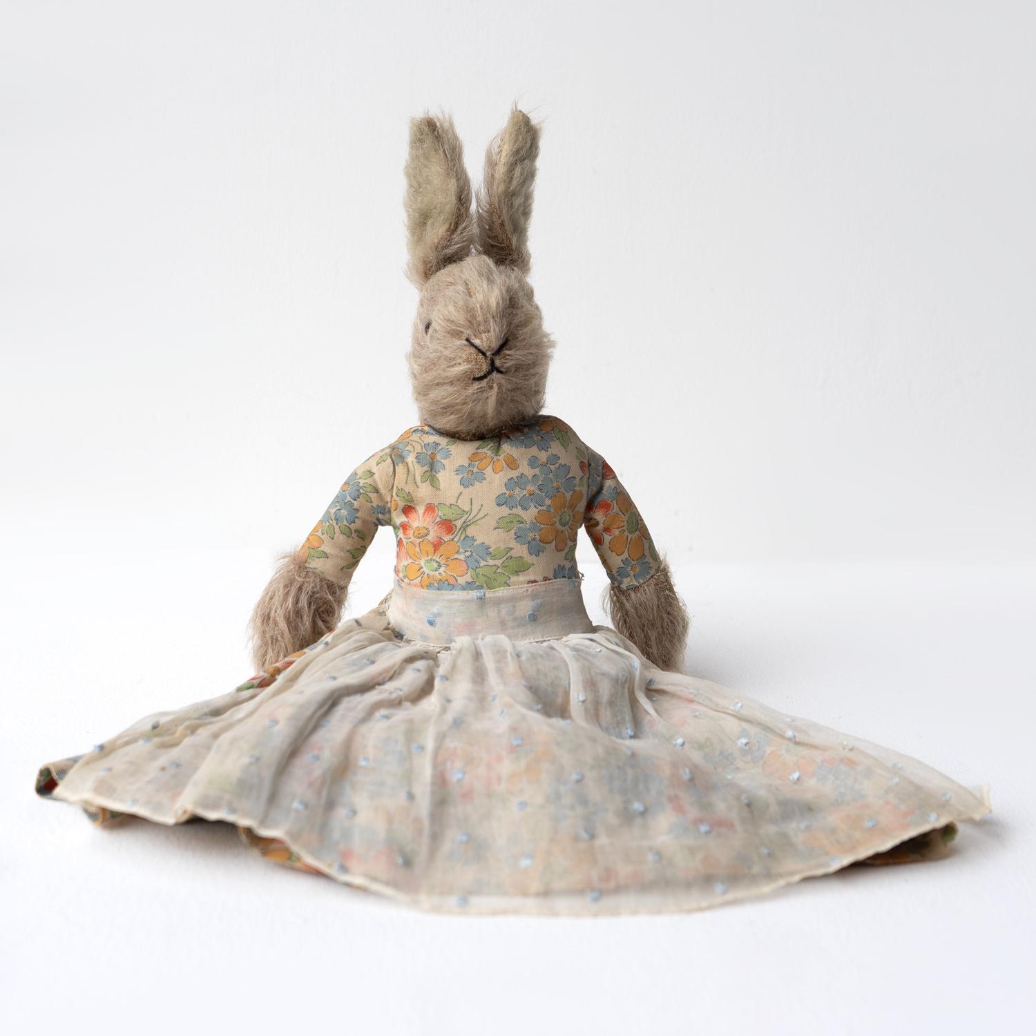 ANTIQUE RABBIT TEDDY

An extremely charming cloth stuffed teddy in the form of a rabbit with grey hair and glass eyes dressed in a pleated floral cotton dress and a voile apron with embroidered polka dots. Underneath the dress, she has white cotton