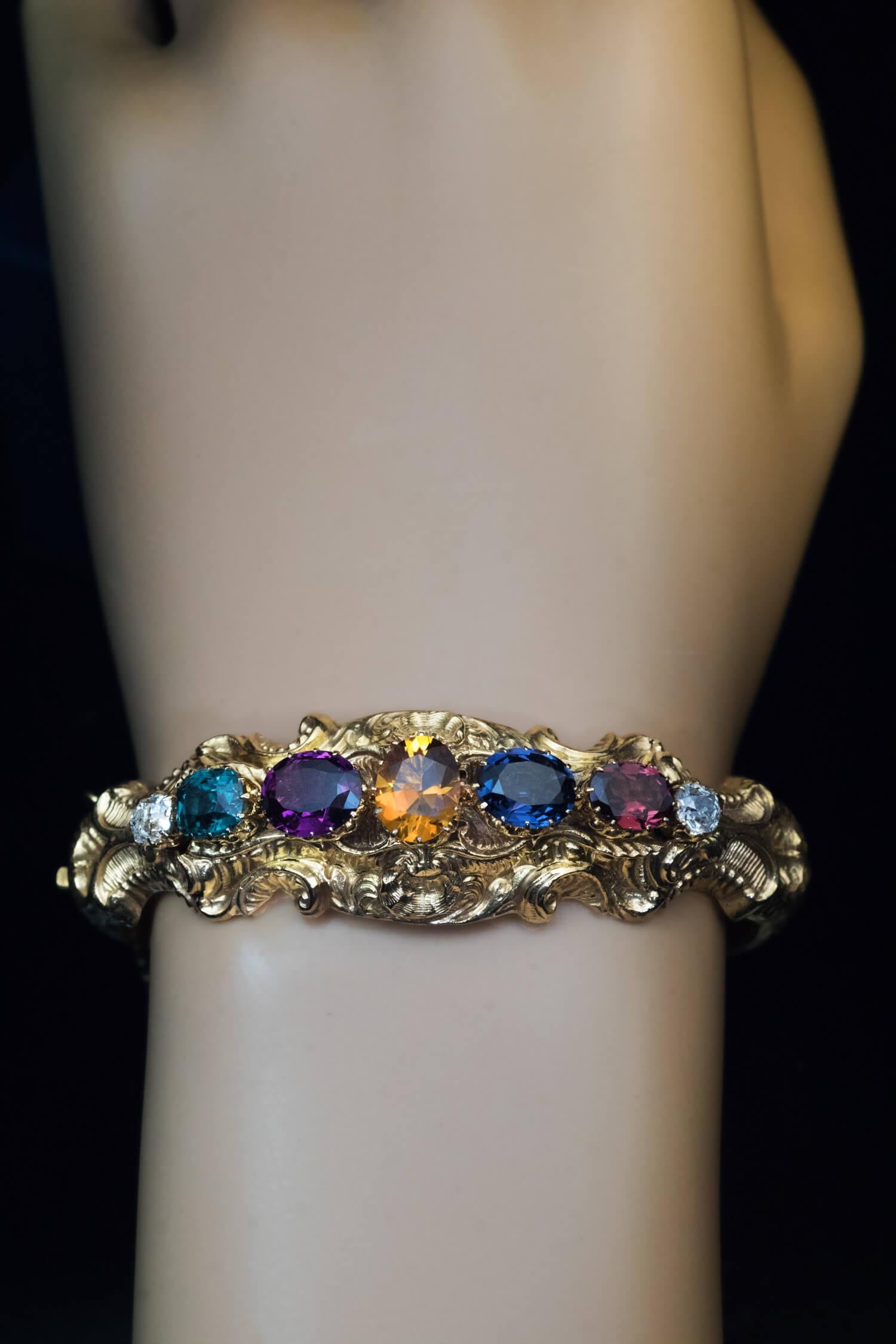 Circa 1850  The hollow 14K yellow gold bangle bracelet is embossed with Rococo style scrolls, foliage and shells. The bracelet is set with two bright white (E color) old mine cut diamonds (approximately 0.60 ct total), a blue spinel, a purple