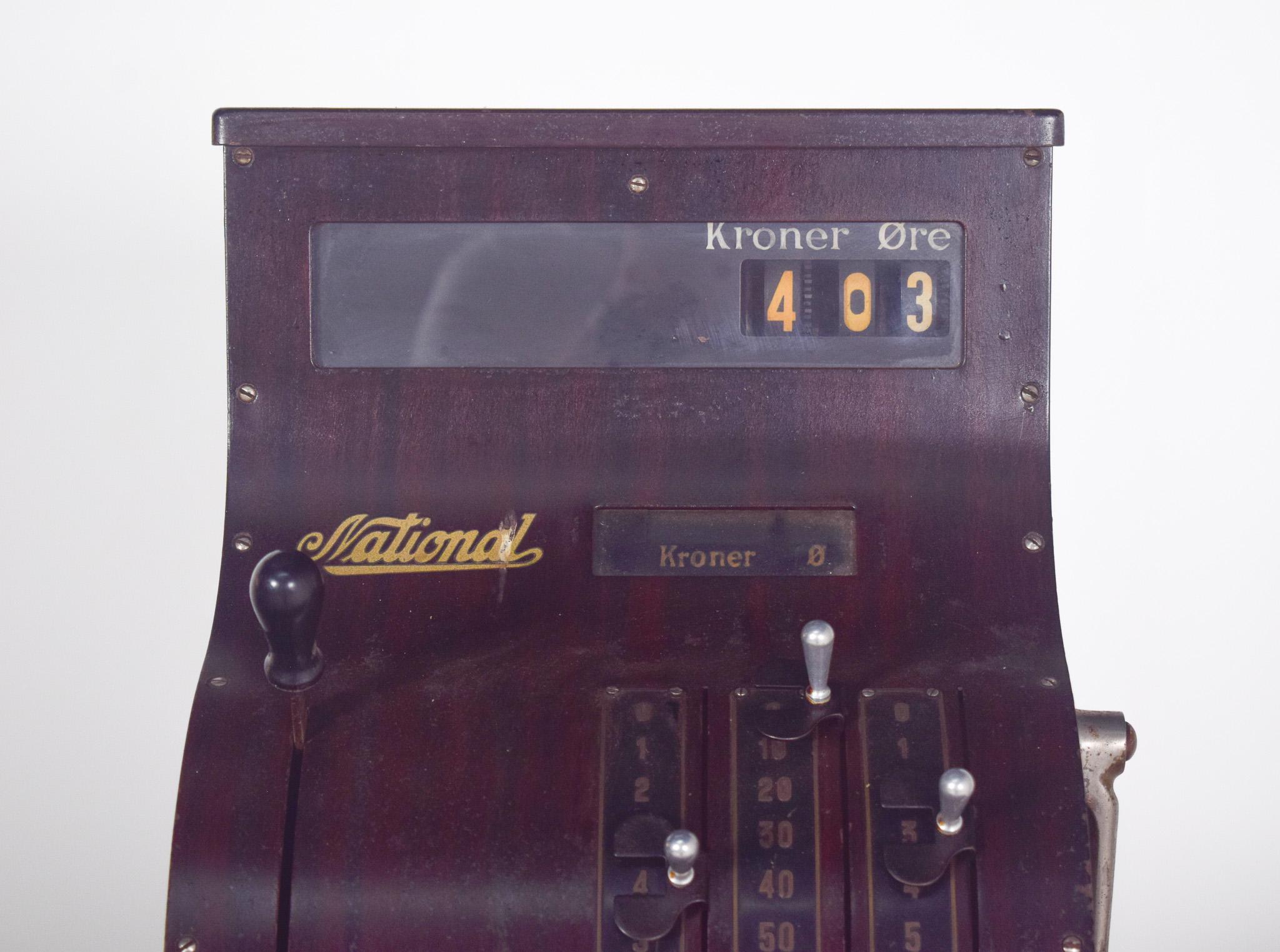 Caisse enregistreuse nationale. Conçu pour les transactions en couronnes danoises (Kroner Øre). Son design classique associe un cadre robuste en bois verni foncé à un mécanisme en métal, reflétant le style des caisses enregistreuses du début du XXe