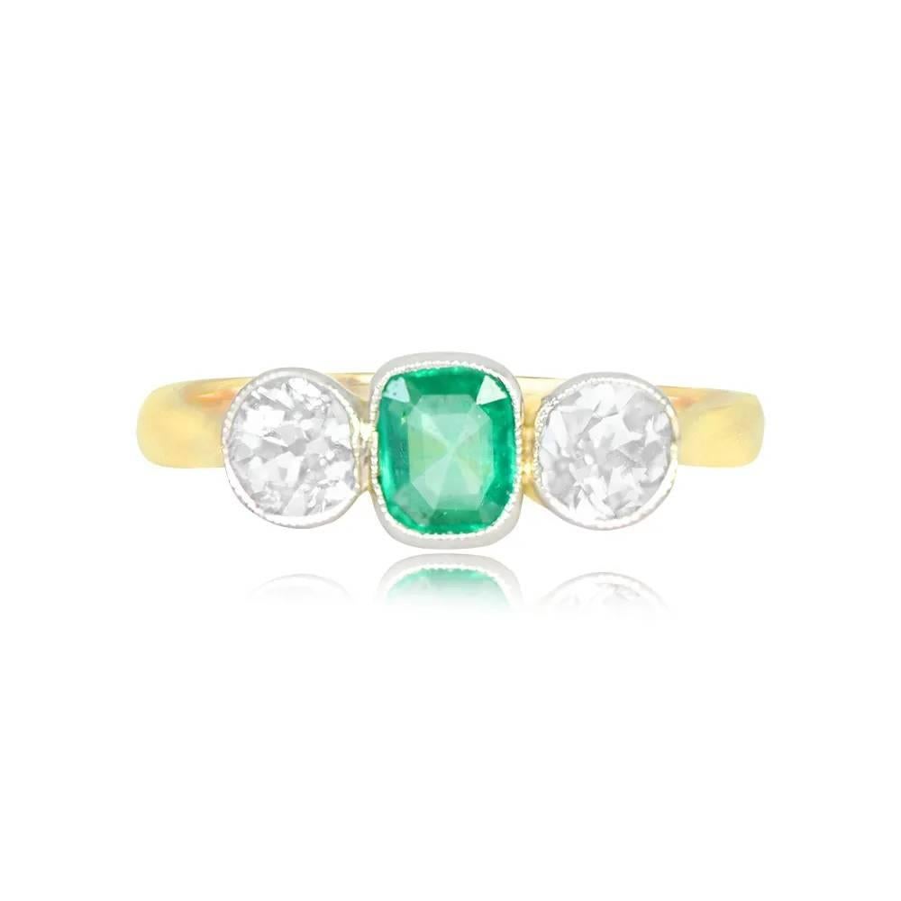 This exquisite antique ring showcases a striking three-stone design. At its center, a natural cushion-cut emerald, elegantly bezel-set, graces the ring, weighing approximately 0.70 carats, radiating a vivid green hue. Flanking the emerald are two