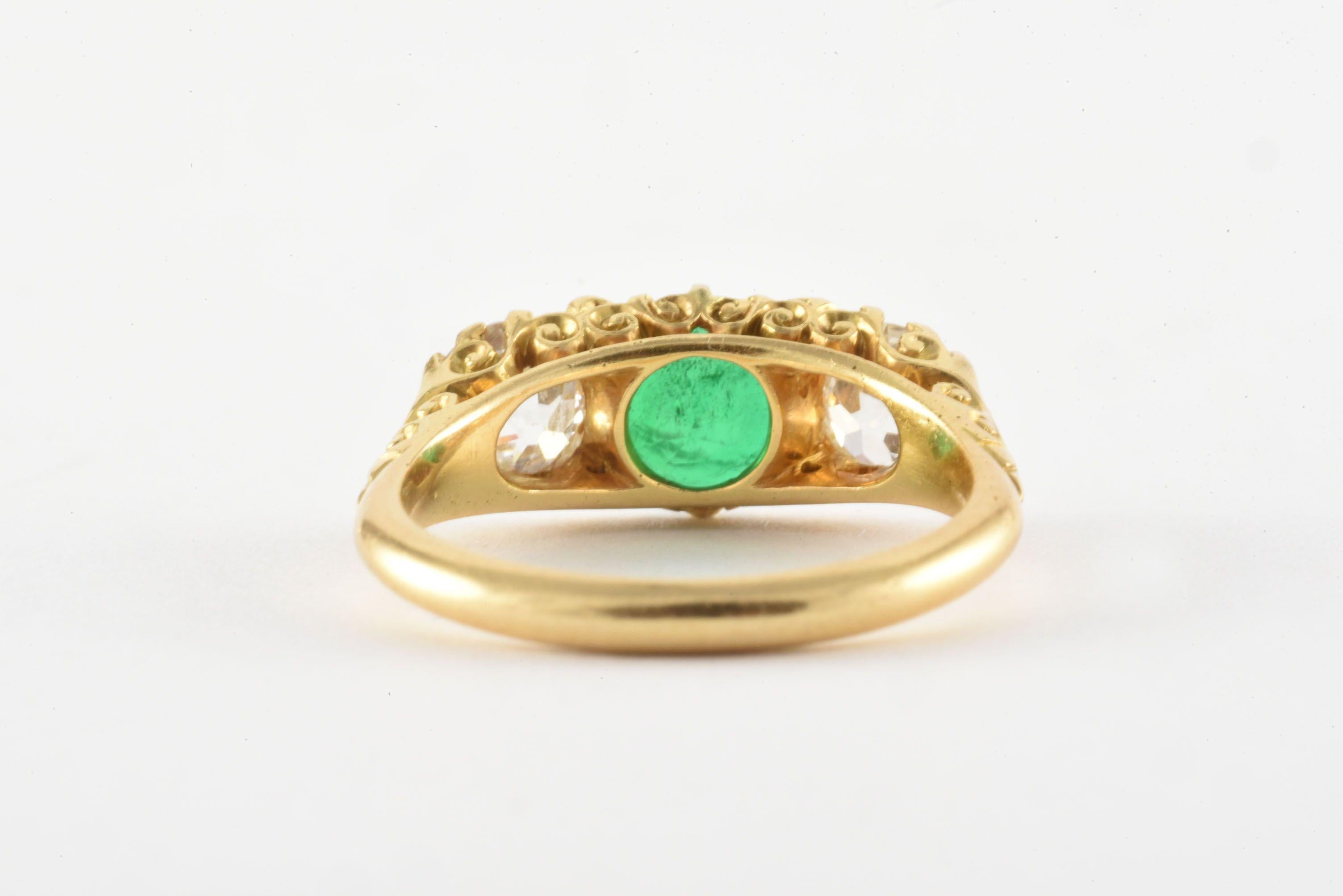 A vibrant natural green round-shaped emerald is flanked by two Old Mine cut diamonds totaling 1.25 carats, F-G color, VS-SI clarity, in this dazzling Victorian Era three-stone band crafted from 18kt yellow gold and accented with scrolling detailed
