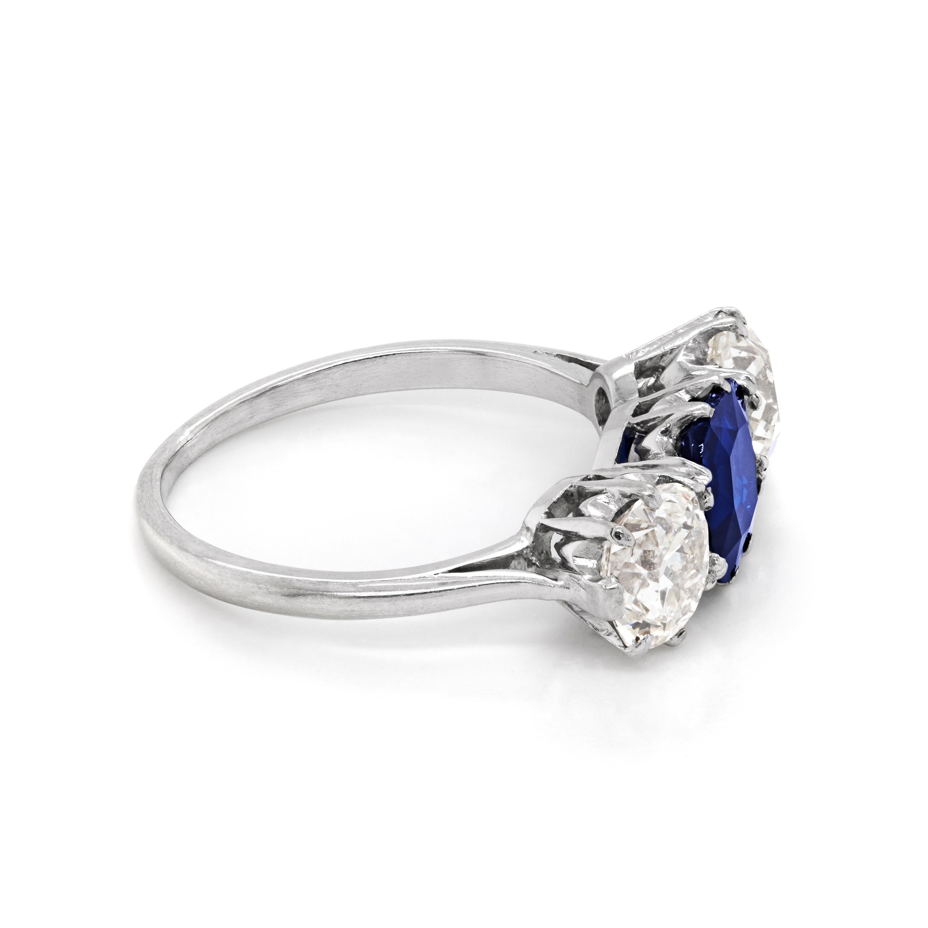 This exquisite three stone ring features a cushion shaped old cut natural unheated transparent blue sapphire mounted in an open back claw setting weighing 1.38 carats. The sapphire is beautifully accompanied by two old cut diamonds with an