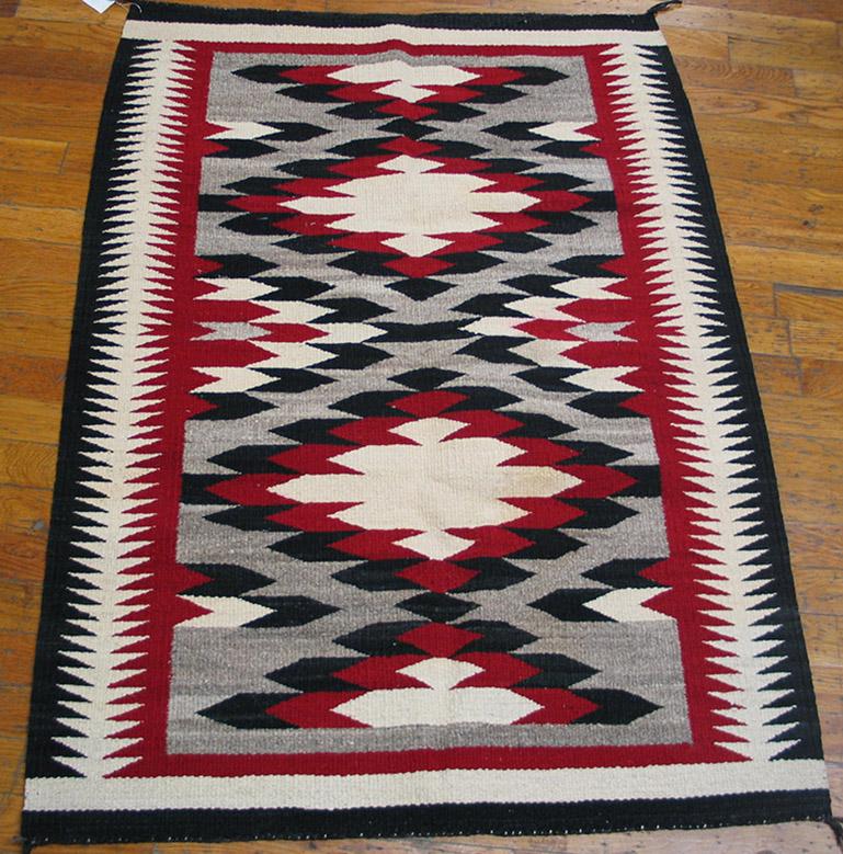 Converging triangle herringbone side borders in ecru, red and black are terminated by solid ecru bands, while the light grey natural wool field shows two complete and four partial lozenge medallions covered with feather patterns in red, black and