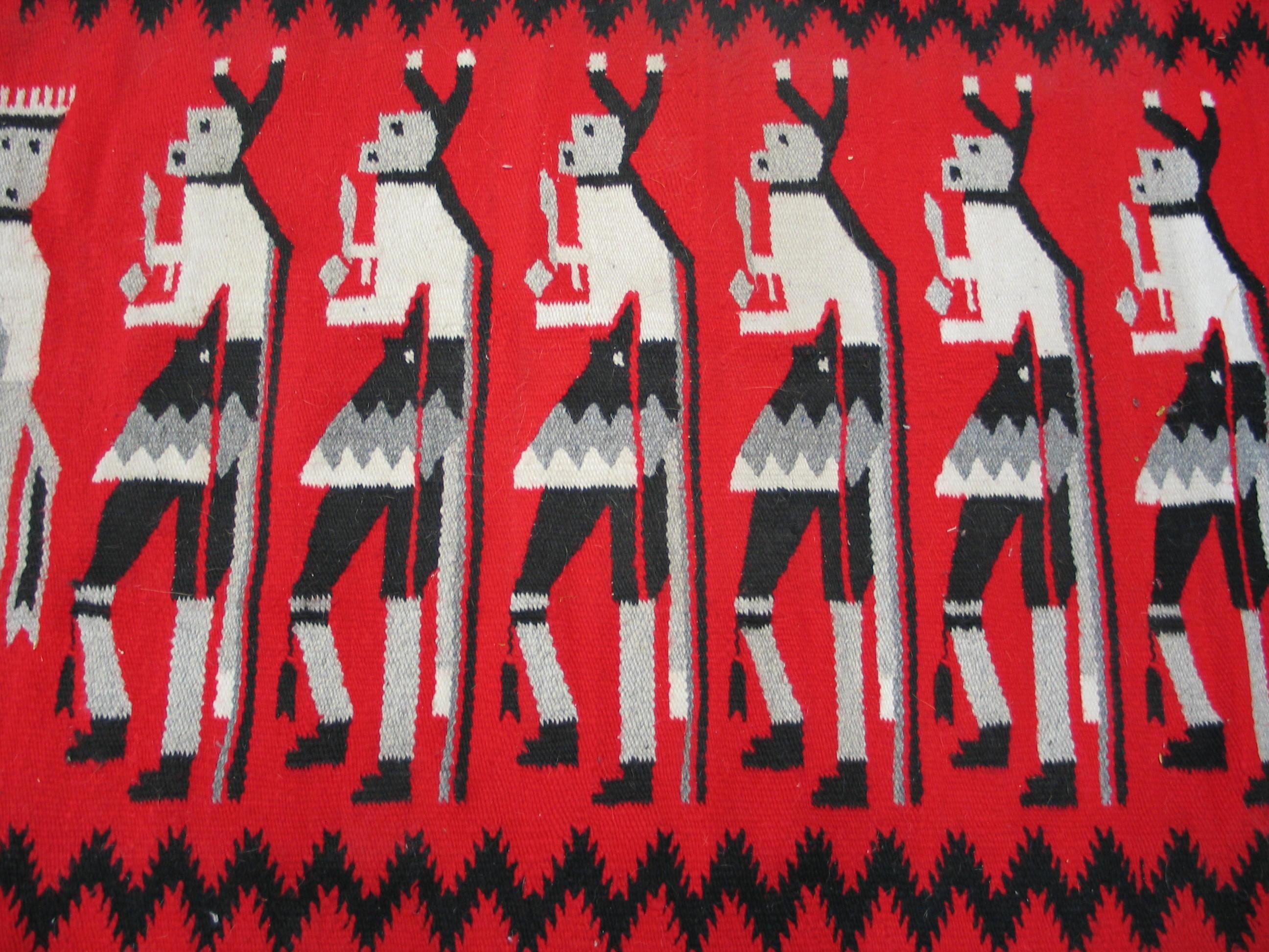 This vintage Yei rug depicts 10 costumed dancers in left profile and two additional end performers, on an unusual red ground. The figures are framed by black barbed zig zags above and below and a surrounding wide off white border with a Caucasian