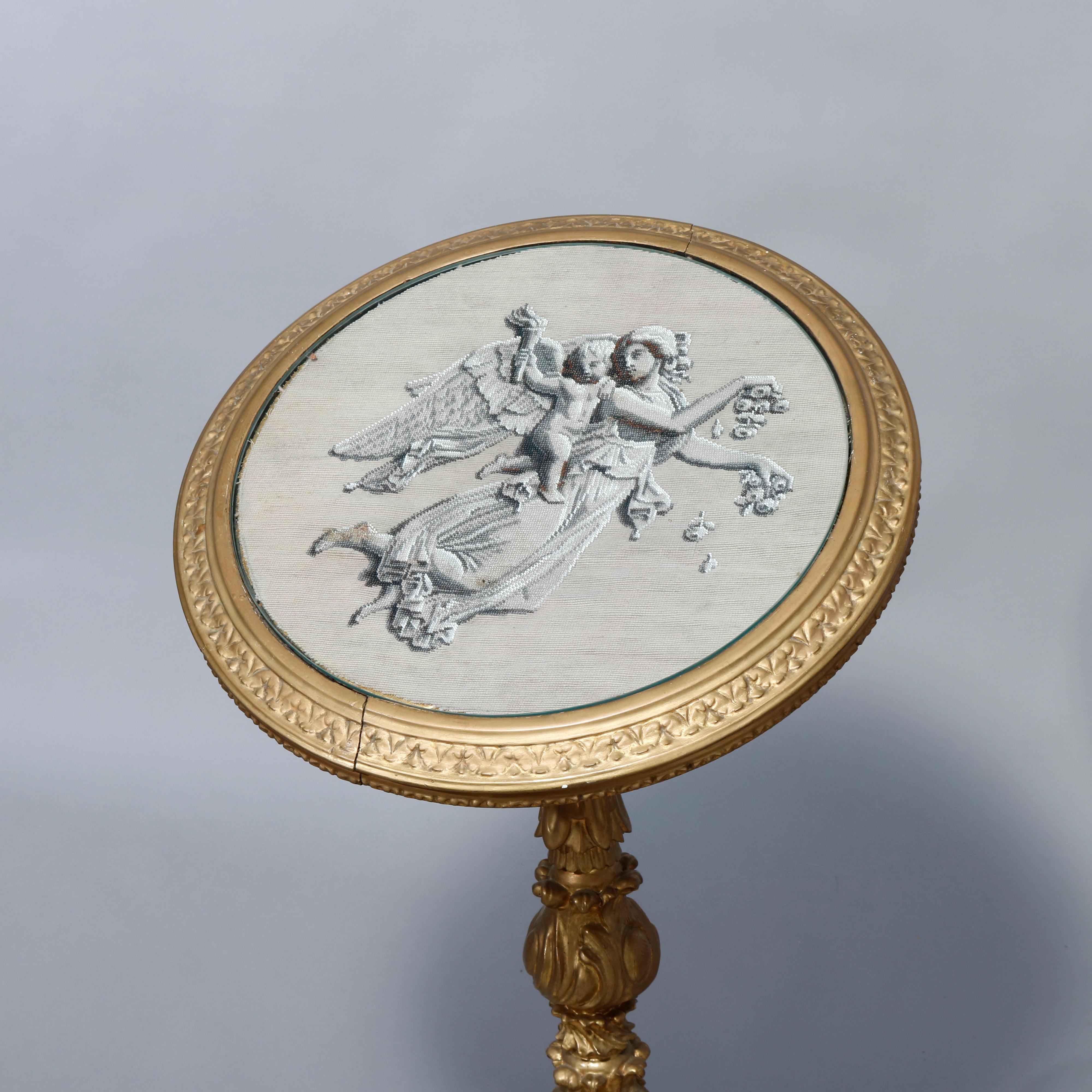 An antique Italian Neoclassical side table offers tilt top with needlepoint inset having scene with angel and cherub seated in giltwood frame with beaded trimming and protective glass raised on foliate carved column with three scrolled gadroon form