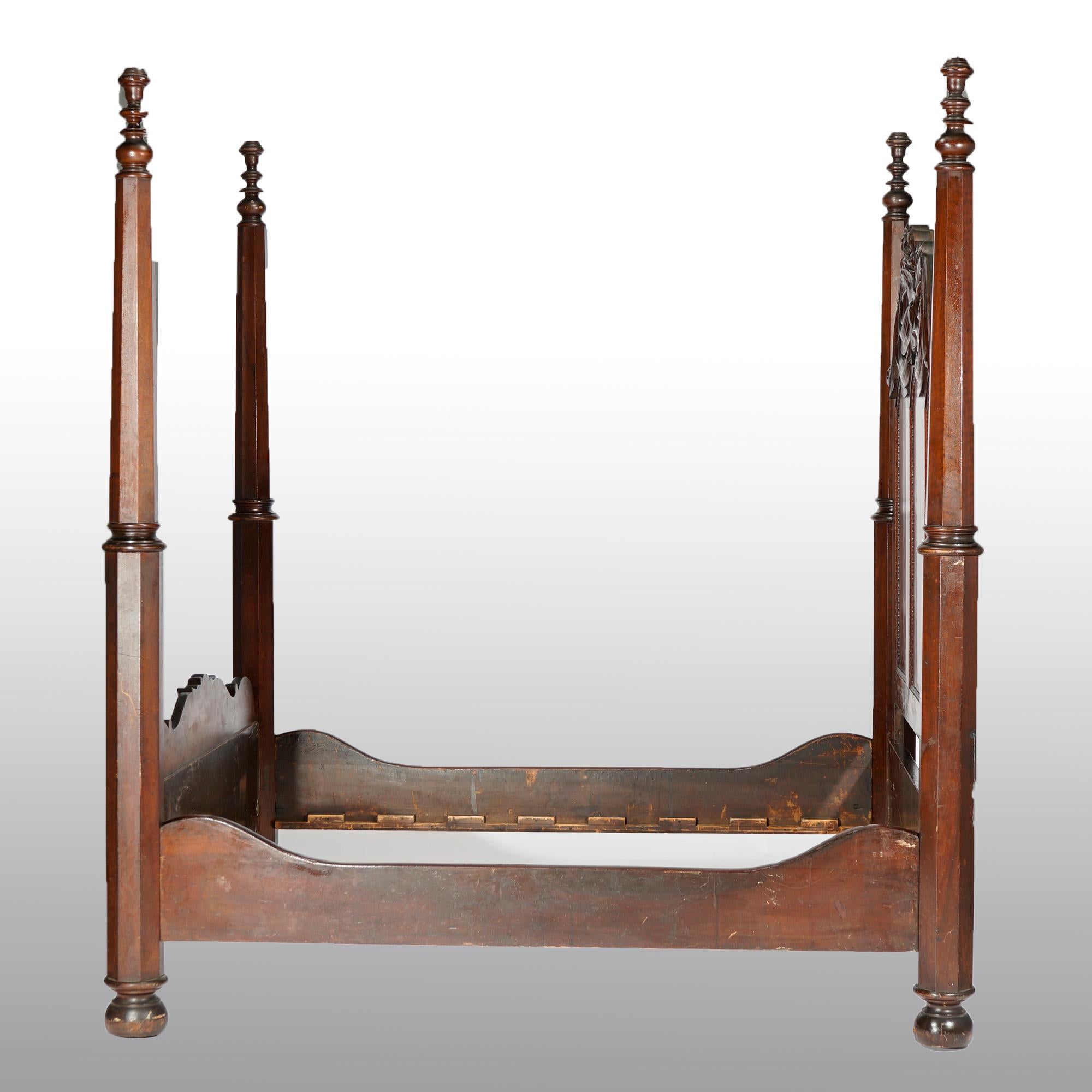 An antique American Empire Greco full size tester bed offers flame mahogany construction with paneled headboard in arch form with carved foliate, floral and shield crest with flanking faceted supports, raised on bun feet, carved decoration