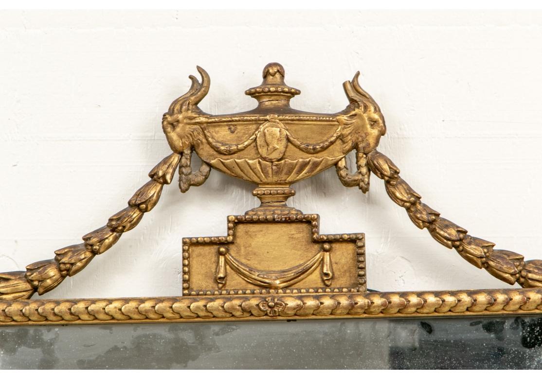 A wide mirror with elaborate crest with center urn decorated with a medallion and goat head and ring handles that support bell flower vines. The urn is raised on a tiered pedestal with a swag. The outer surround with overlapping discs and goat heads