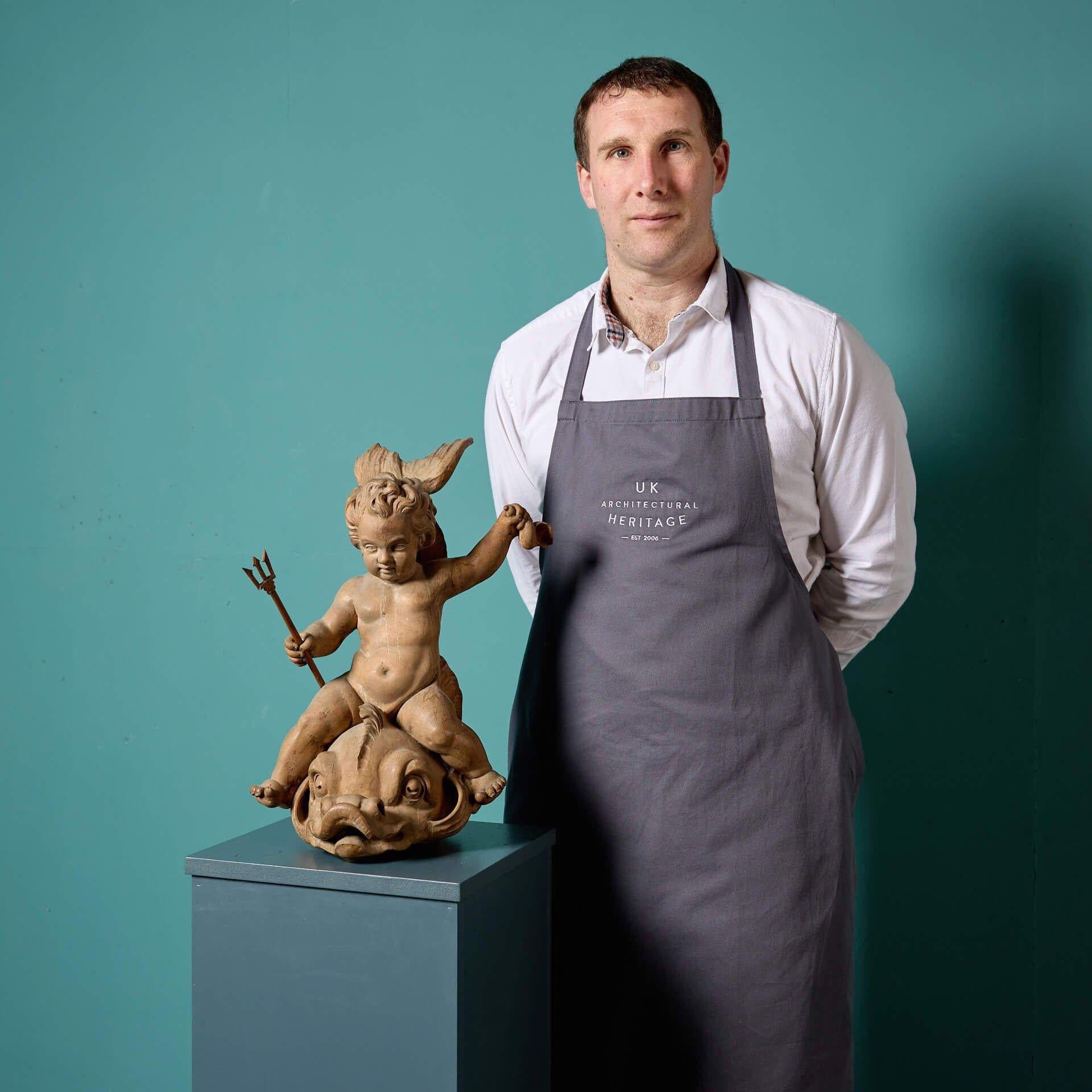 Ein prächtiger, kunstvoll geschnitzter Putto aus dem frühen 19. Jahrhundert, der auf einem stilisierten Delphin sitzt. Die aus Buchenholz gefertigte neoklassizistische Skulptur stellt einen detailreichen Putto dar, der in der einen Hand eine