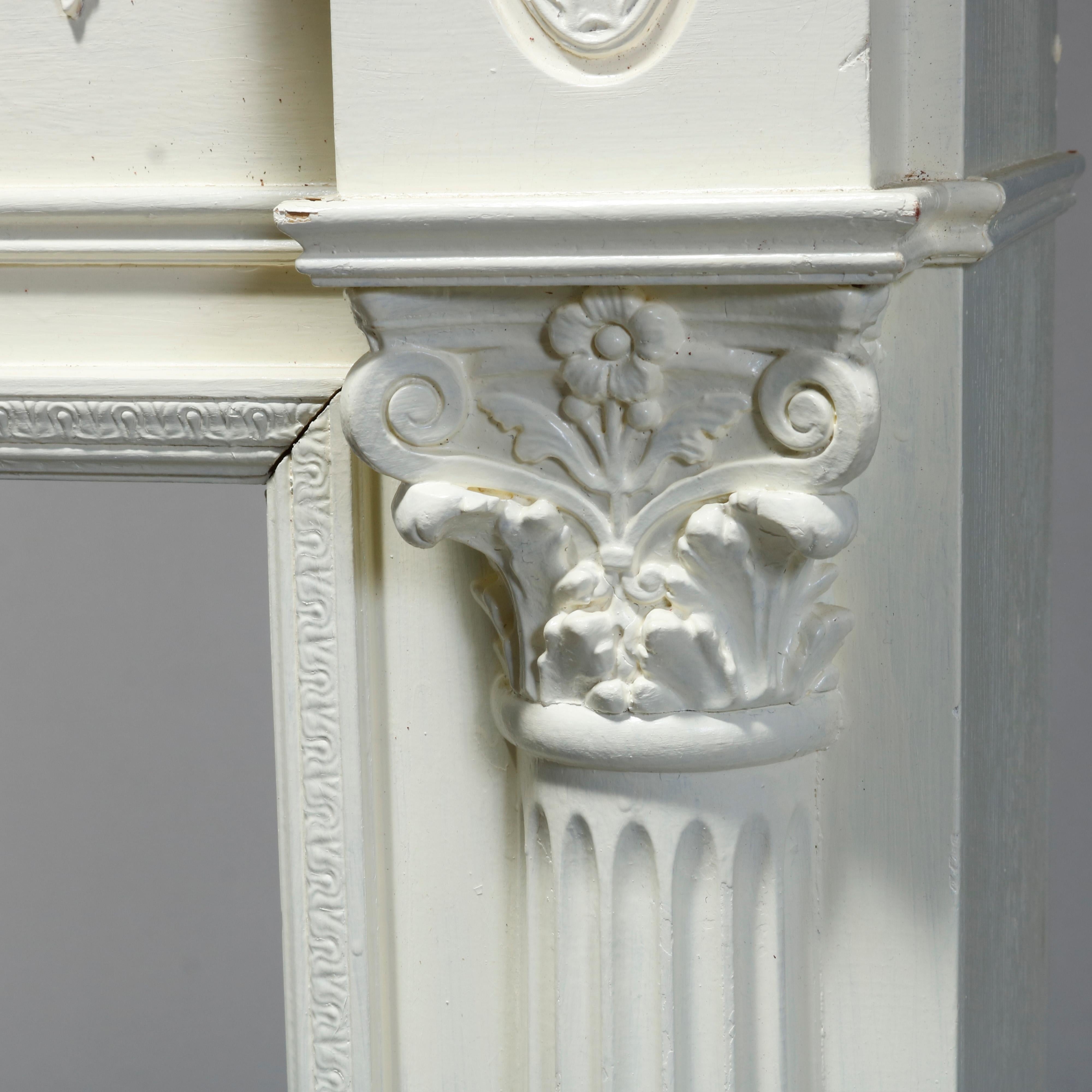 An antique neoclassical architectural fireplace mantle offers wood construction with frieze having applied repeating wreath and swag garland elements with central urn and flanking rosettes over Corinthian column supports, painted white, 20th