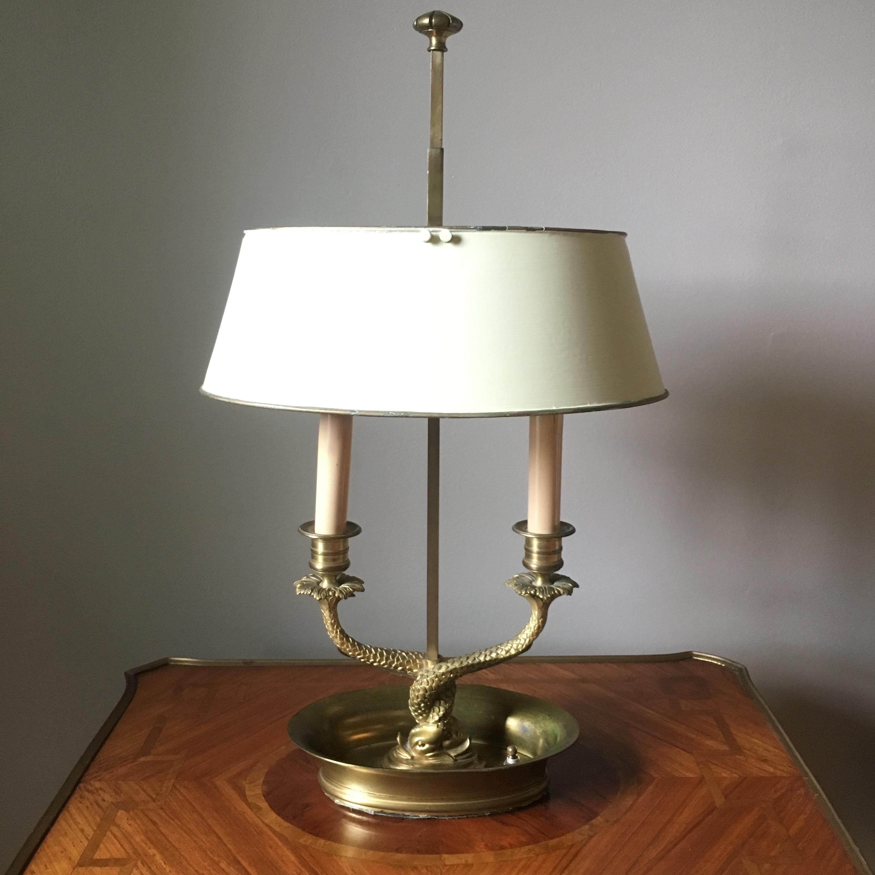 A stunning Neoclassical style brass bouillotte lamp with twin dolphins at the base and a cream colored tole shade. 

Early 20th Century

Dimensions: 12ʺW × 8ʺD × 21ʺH

Good vintage condition. Minor surface wear expected with age and use. Lamp powers