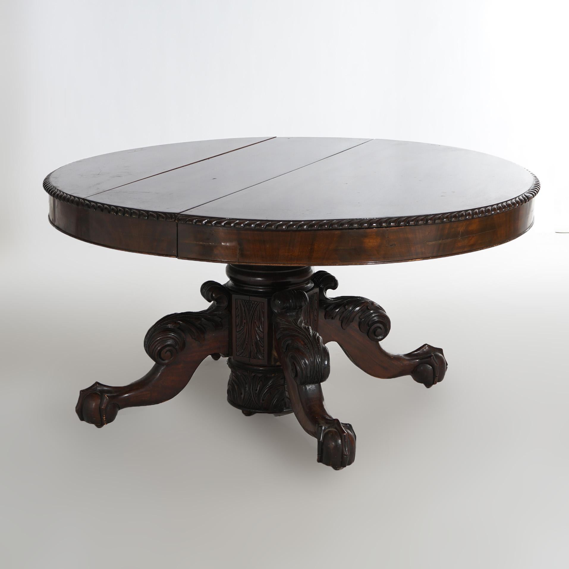 An antique Neoclassical Greco American Empire extension dining table offers flame mahogany construction in round form with carved gadroon trimming over stylized urn form pedestal and raised on foliate carved cabriole legs terminating in claw and
