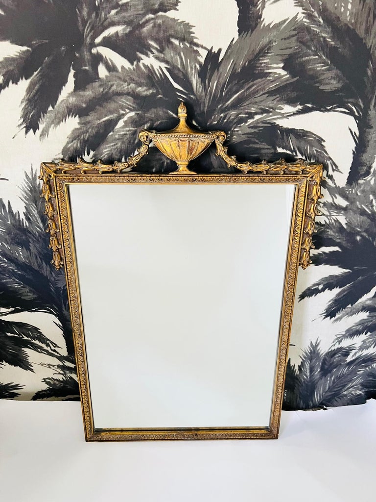 French antique Neoclassical mirror with gold leaf frame. The hand carved mirror features an urn pediment with stylized bell flowers draped over the sides of the frame. Hand carved laurel motifs decorate the interior borders. The scale makes for a