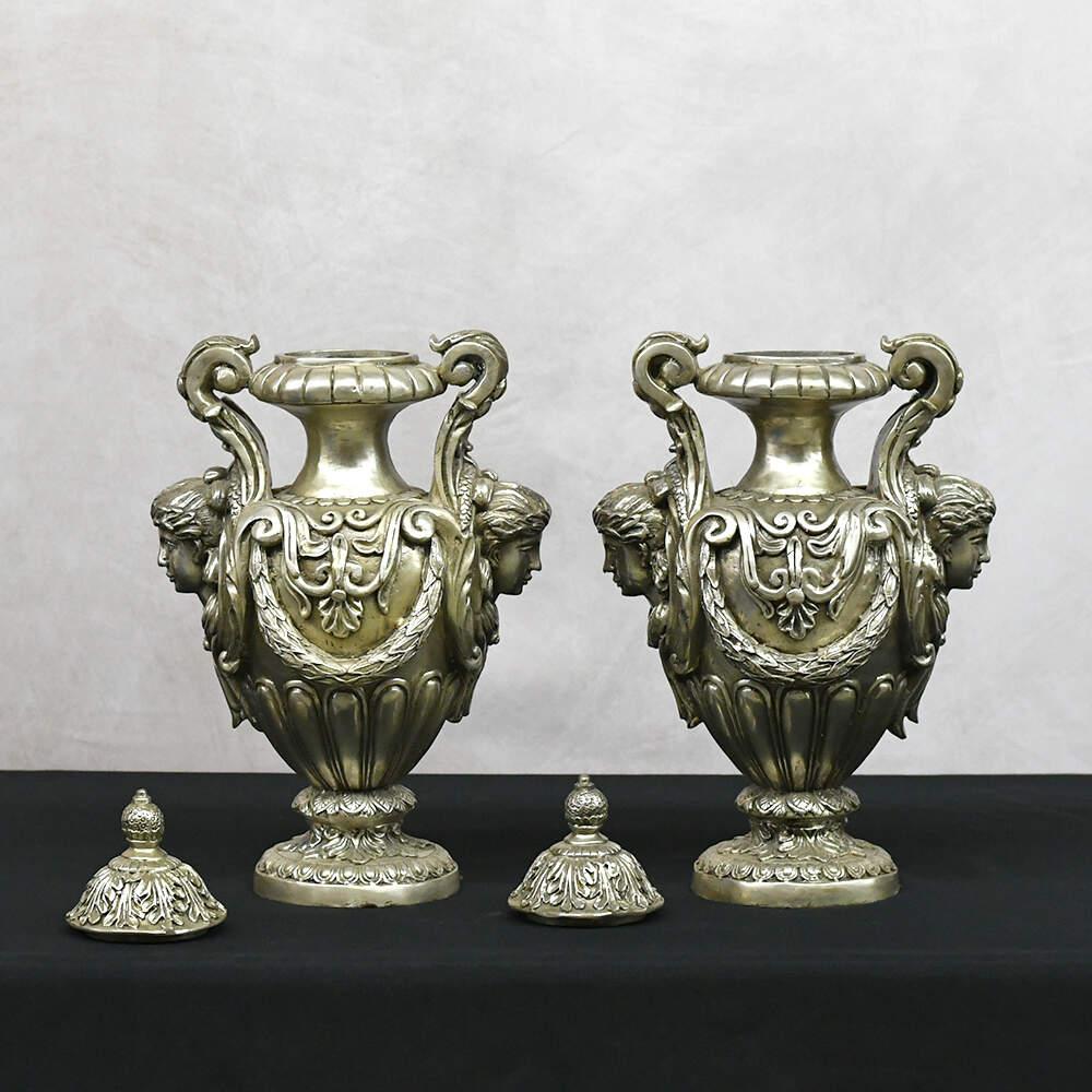 This commanding pair of Neoclassical Maiden's Visage Covered Urns brings classical grandeur into the present with an unmistakable presence. Cast in a lustrous metallic finish, each urn is adorned with finely rendered maiden's faces that emerge from