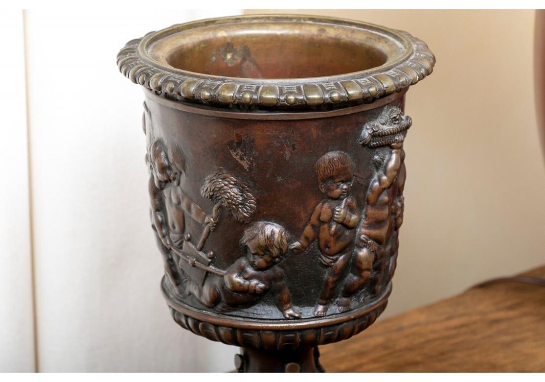 A footed urn with relief decoration of putti in high relief cavorting in a landscape as gardeners. They carry a planter with a tree, basket and garlands of flowers. An urn on a pedestal appears in the background. The foot with leaf and berry trim
