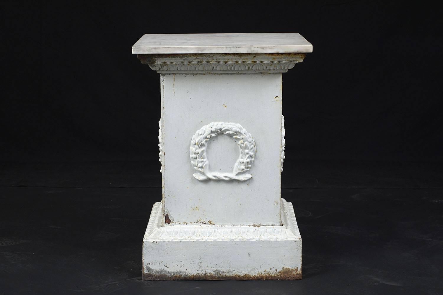 This 1900s neoclassical style pedestal is made of iron painted an off-white color with natural oxidization and rust accents. The pedestal is adorned with egg and drop bands along the top and bottom and a garland wreath on every side. The top of the