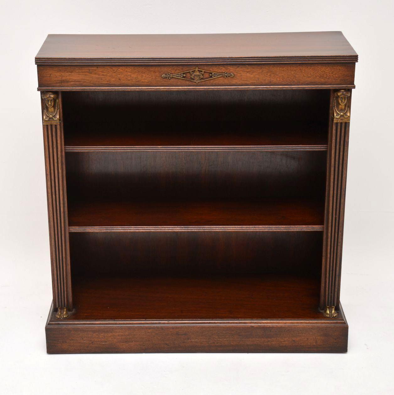 Small antique neoclassical Empire style mahogany open bookcase in excellent condition and dating from circa 1930s period. It has a reeded top edge and reeded tapered side columns, topped with gilt bronze faces with feet at the base. The shelves have
