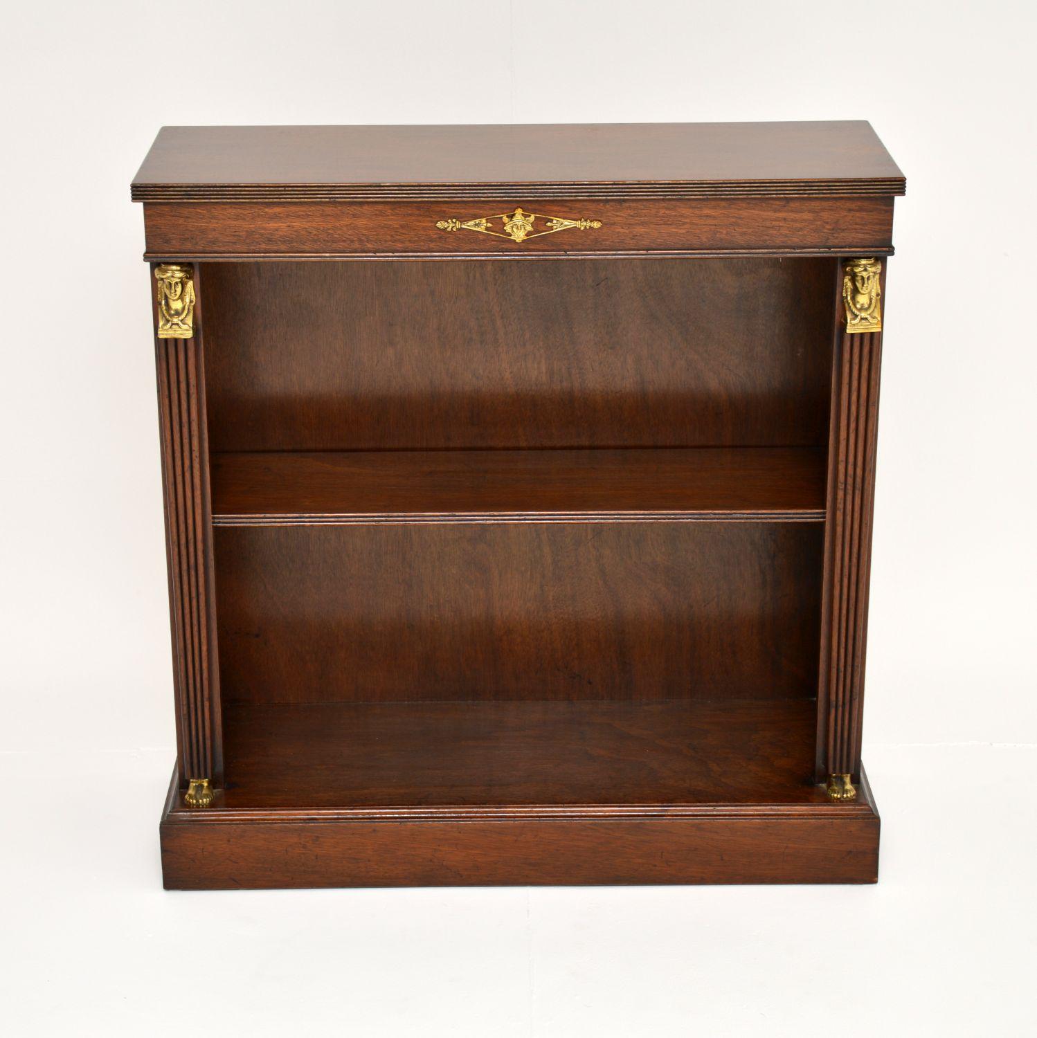 A lovely antique neoclassical Empire style wood open bookcase, dating from around the 1930-50’s period.

It has a reeded top edge and reeded tapered side columns, topped with gilt bronze faces with feet at the base. The shelf has a reeded front &