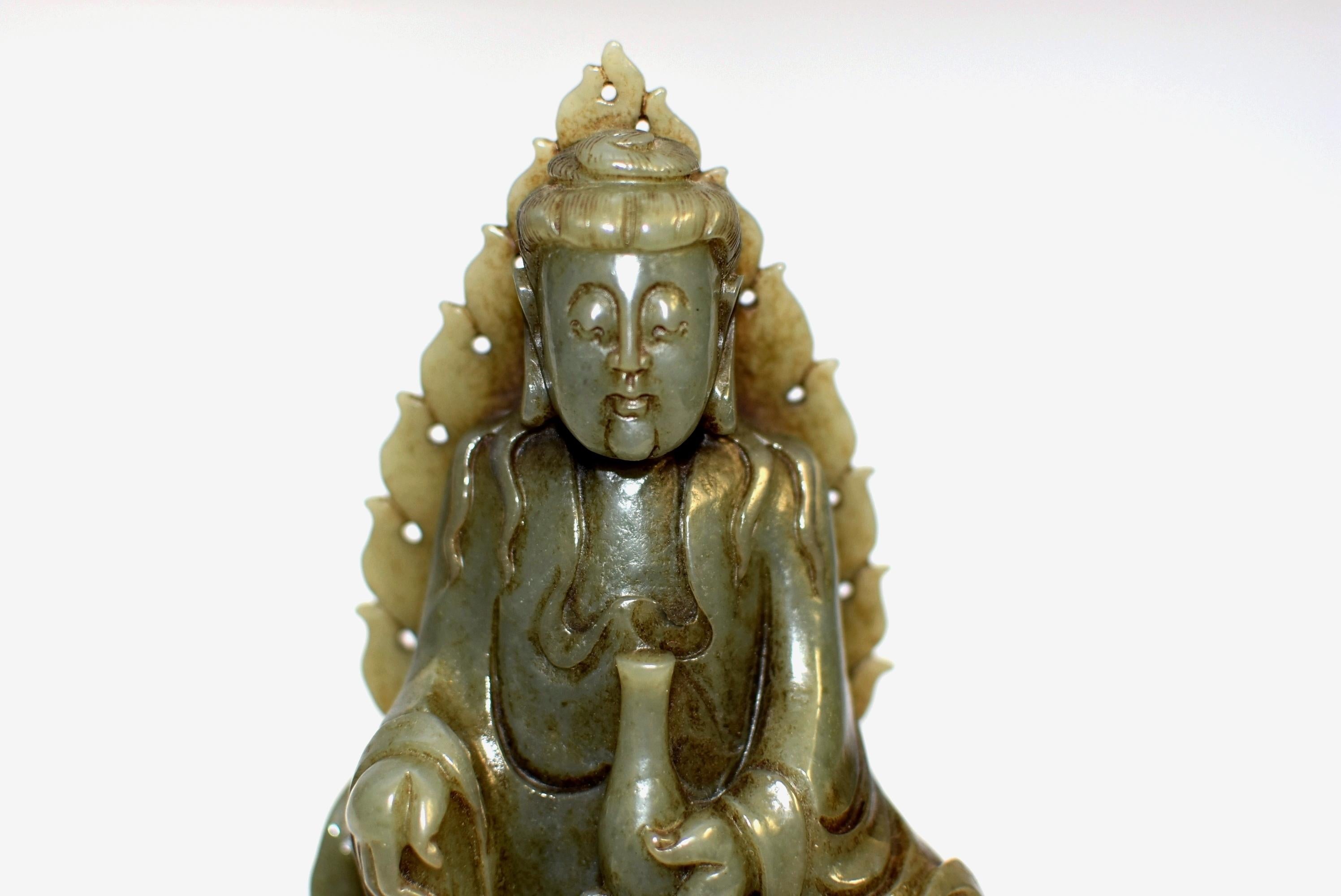 A Qing dynasty, hand carved, lustrous, solid natural nephrite green jade Buddha. Carved seated on a rock against flame-shaped mandorla, the broad-faced figure rendered with a serene expression by downcast eyes beneath high arched brows above pursed