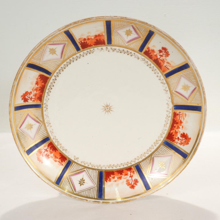 A fine antique English porcelain dish or plate.

By New Hall.

With an eye-catching gilt rim that alternates between panels of iron-red underglaze countryside and gilt geometric and floral devices with bright blue painted accents. There is a