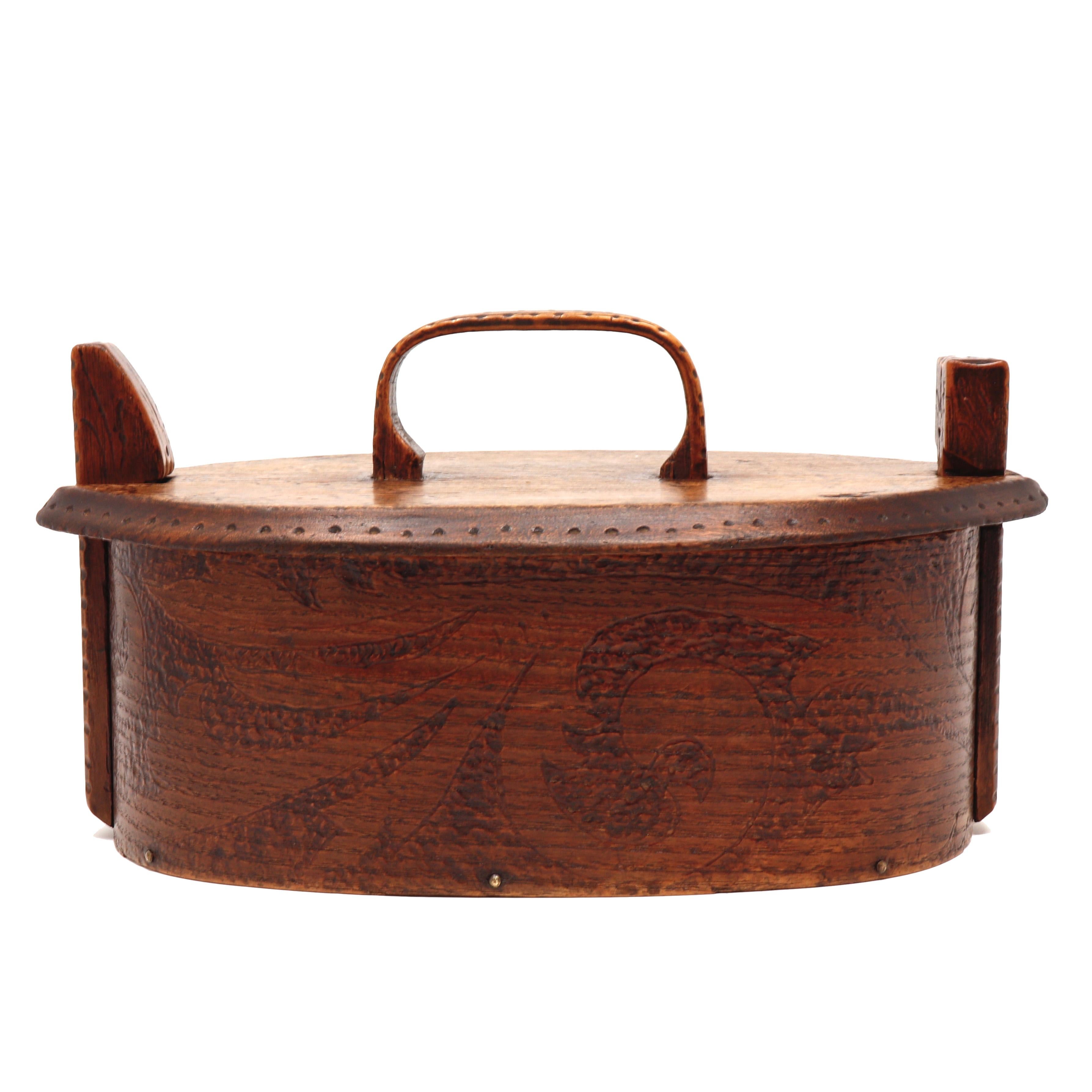 A lidded wooden basket, the sides from one piece of thin steamed wood tacked together with brass brads at the centre of one side and beveled decorative seam, flat wood bottom and flat wooden top and handle that locks into place by the design of the