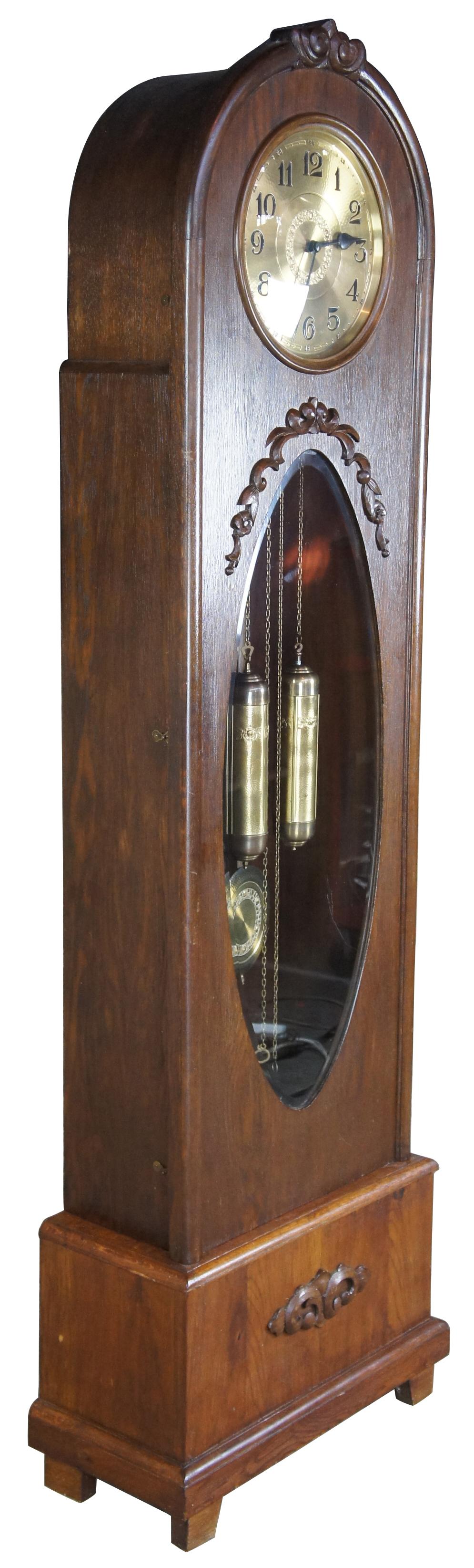 Art Deco German grandfather clock by Kieninger. This tall case clock has an oak wooden case with a round bonnet, and a plinth base featuring an appliqued foliate motif rising on squared feet. The front of the clock has an oval glass door with arched