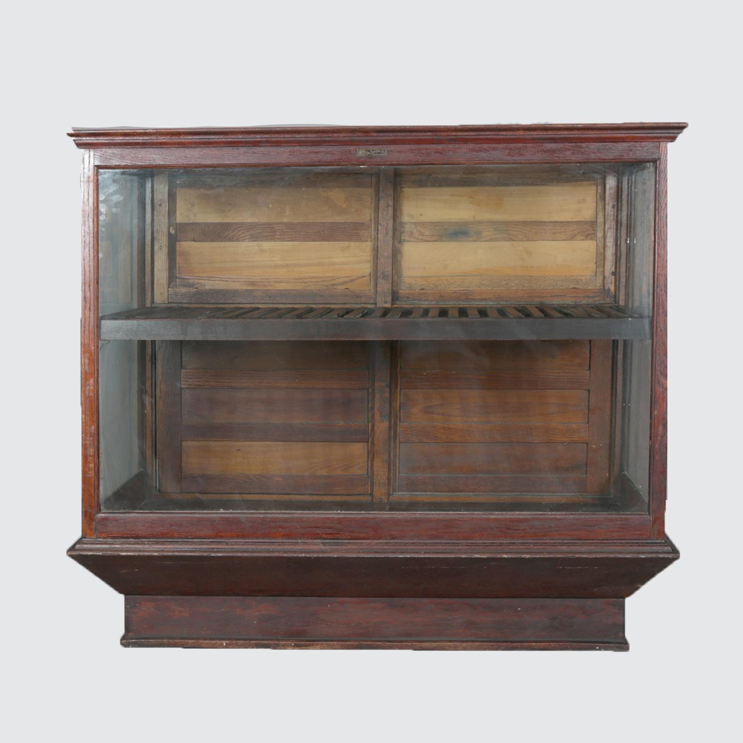 An antique Sun Manufacturing Company country store display case features oak construction with glass front, top and sides with back having upper and lower sliding doors opening to interior with slat shelf, raised on faceted plinth, maker plaque on