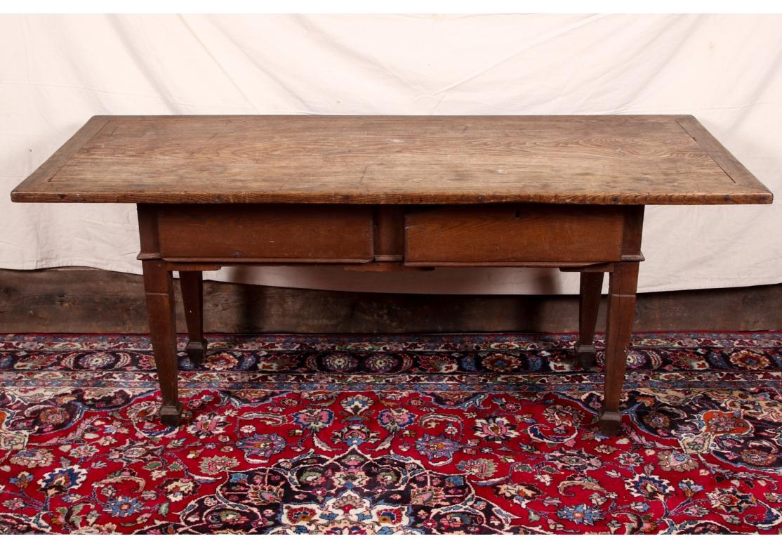 A very old and solid work table which shows its age to great advantage. With a long banded overhanging top, two apron drawers (one with an escutcheon, lacking the key), and carved frame below the apron. Raised on square legs with spade feet. Lacking