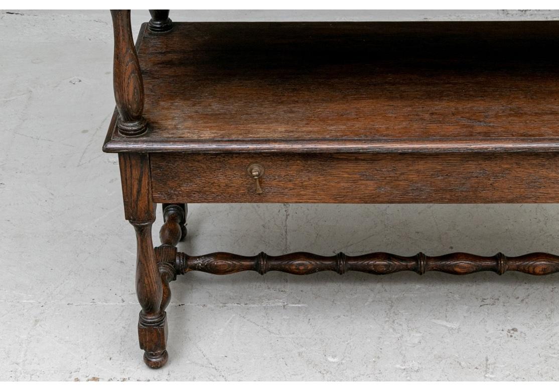 An Antique Tiered Console in an unusual open form. The oak console table with a single long frieze drawer on both the top and lower tier with brass drop pulls. Turned baluster supports and legs with elaborately turned H stretcher and ball feet.