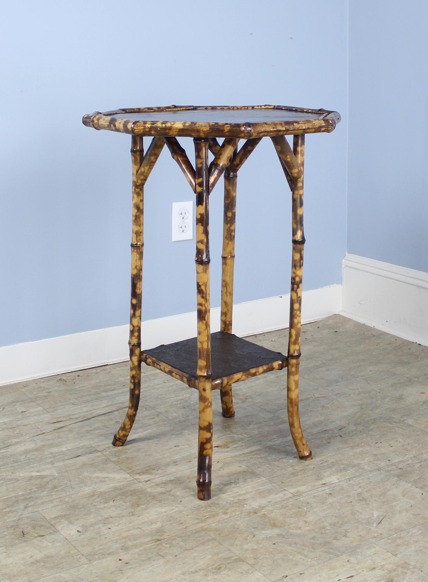 An antique English bamboo side table, with flared legs and an unusual octagonal top accented with a chinoiserie lacquered top with a floral and wildlife motif. The bottom shelf is covered in stylish pressed leather. The bamboo, vividly painted, is