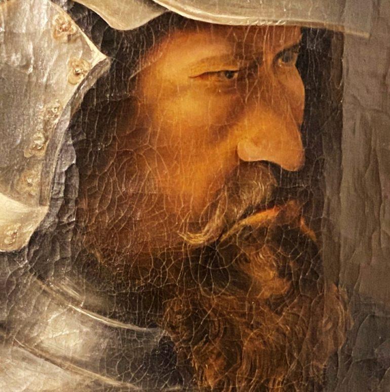 A framed oil-on-canvas portrait of a fierce bearded conquistador wearing armour and a helmet.


