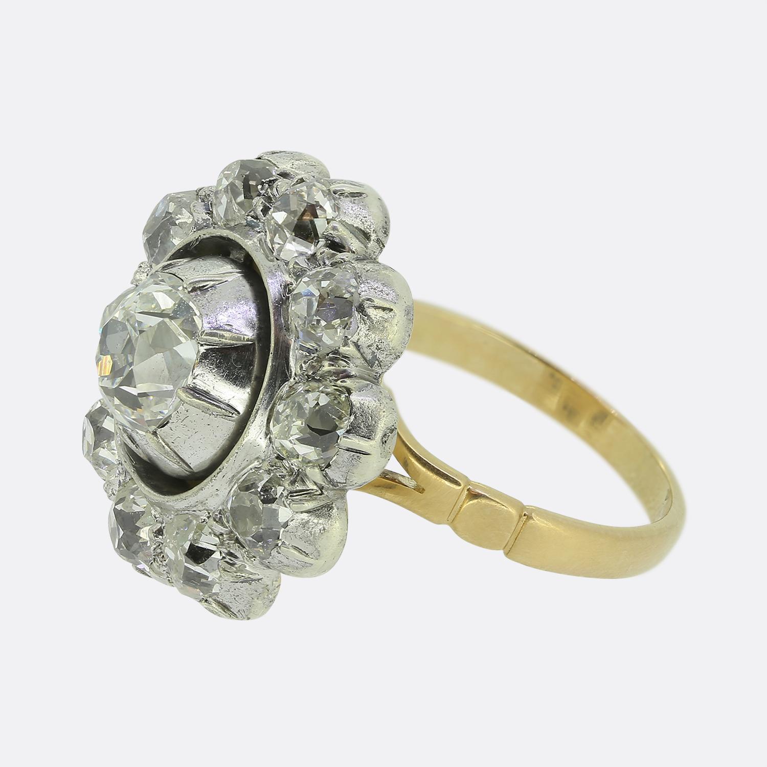 Hier haben wir einen exquisiten Clusterring mit einem antiken Diamanten im Kissenschliff in der Mitte, der einen fesselnden weißen Farbton ausstrahlt. Der Hauptstein ist in einer eleganten Zange gefasst und von einem Kranz kleinerer Diamanten im