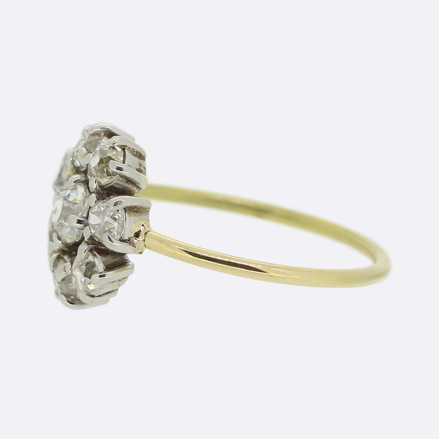 Hier haben wir einen schönen Diamant-Cluster-Ring. In der Mitte dieser offenen Fläche finden wir einen einzelnen klobigen, rund facettierten Diamanten mit altem Minenschliff. Dieser Hauptstein ist von einer kreisförmigen Anordnung von weiteren alten