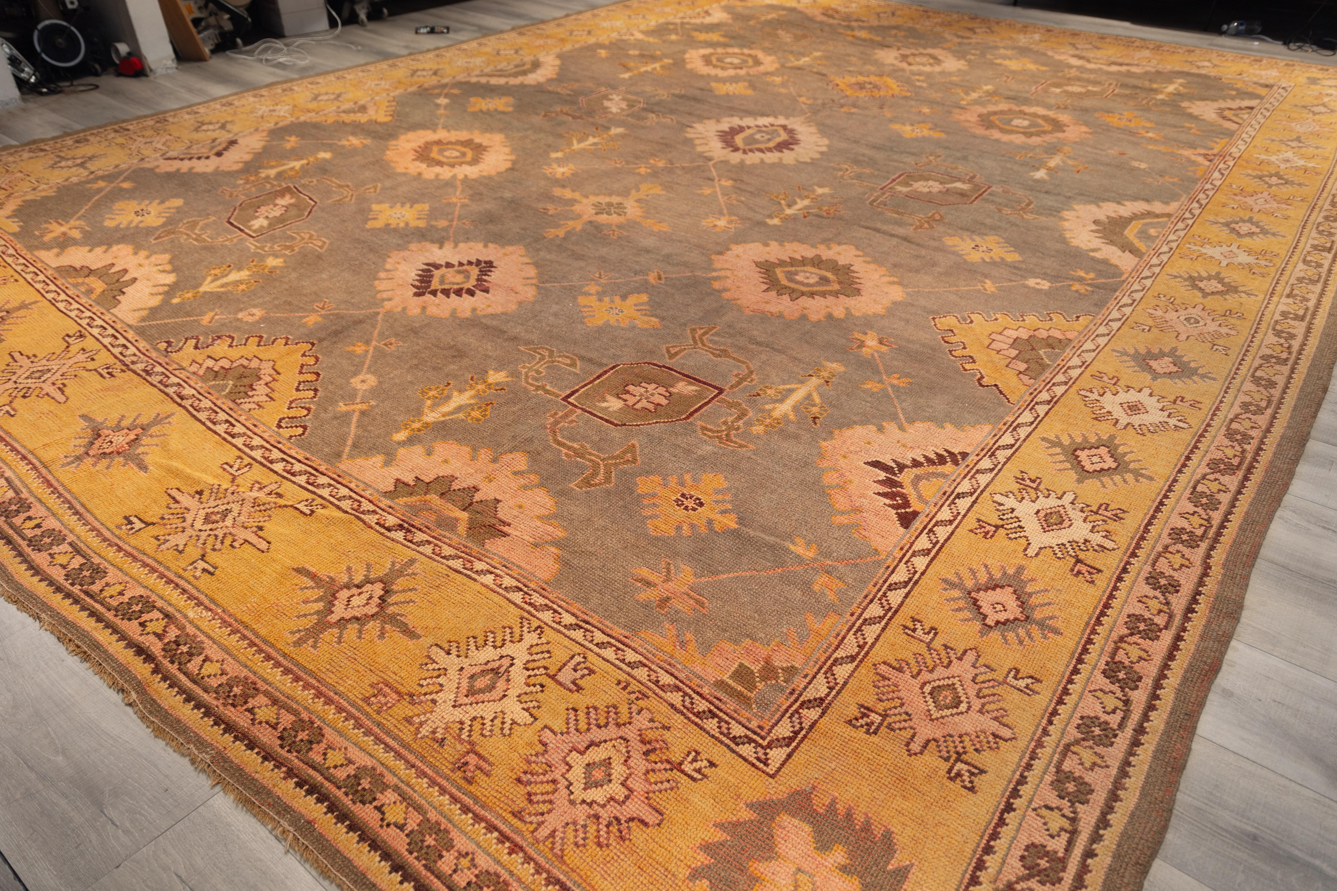 Oushak rug features a soft Olive-brown field with a scattered pattern of large medallions, floral rosettes, and geometric motifs in light peach, gold, and muted burgundy. The border in warm yellow carries a repeating star-like design, tying the