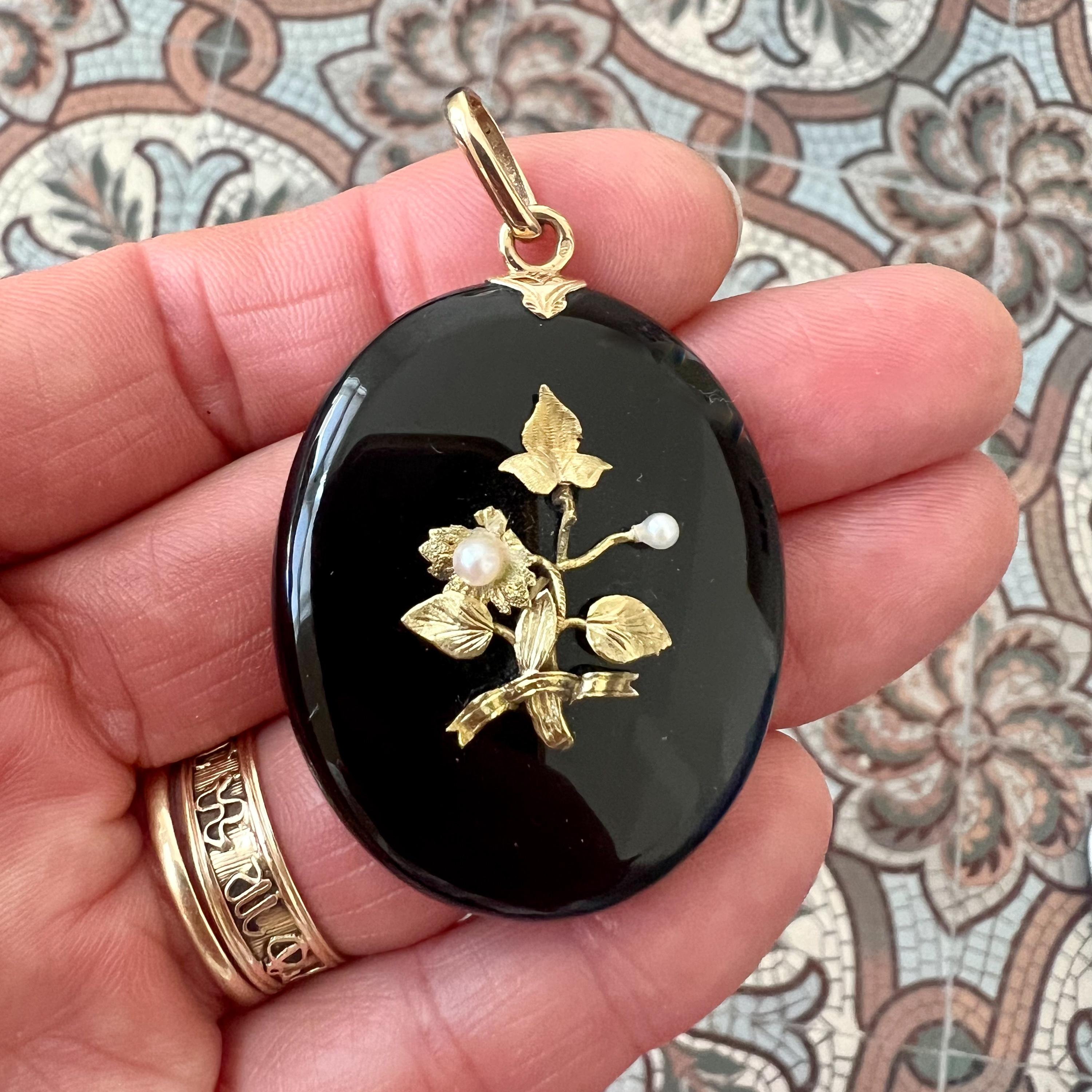Questo bellissimo ciondolo antico a forma di medaglione ovale è finemente realizzato in onice e impreziosito da un'applicazione floreale in oro con due perle coltivate. Sul retro, il pezzo si apre per rivelare uno scomparto per le foto, che contiene