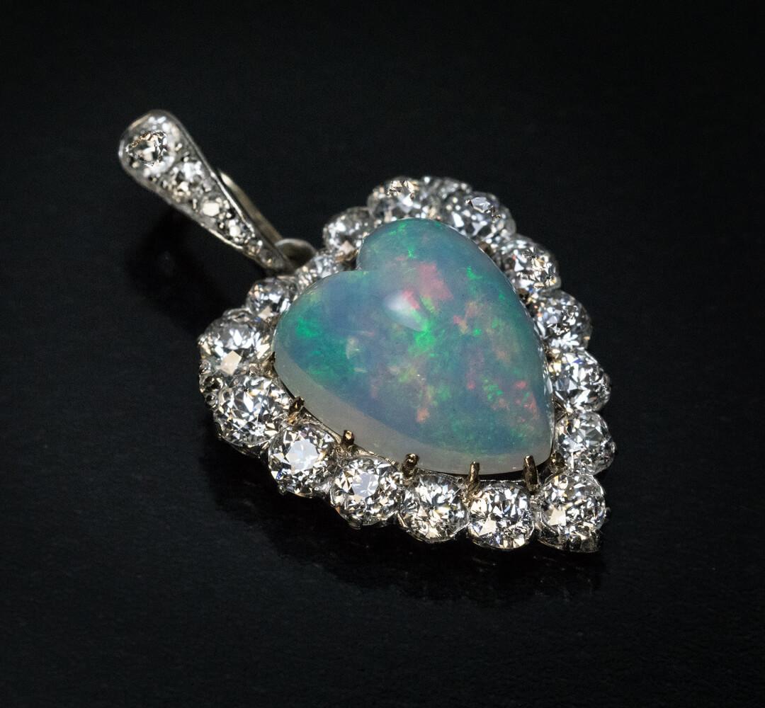 Circa 1910
The pendant features a heart-shaped opal of an excellent greenish blue color with splashes of pink. The opal is framed by bright white old European cut diamonds. The diamonds are set in platinum over 14K gold. The bail is set with