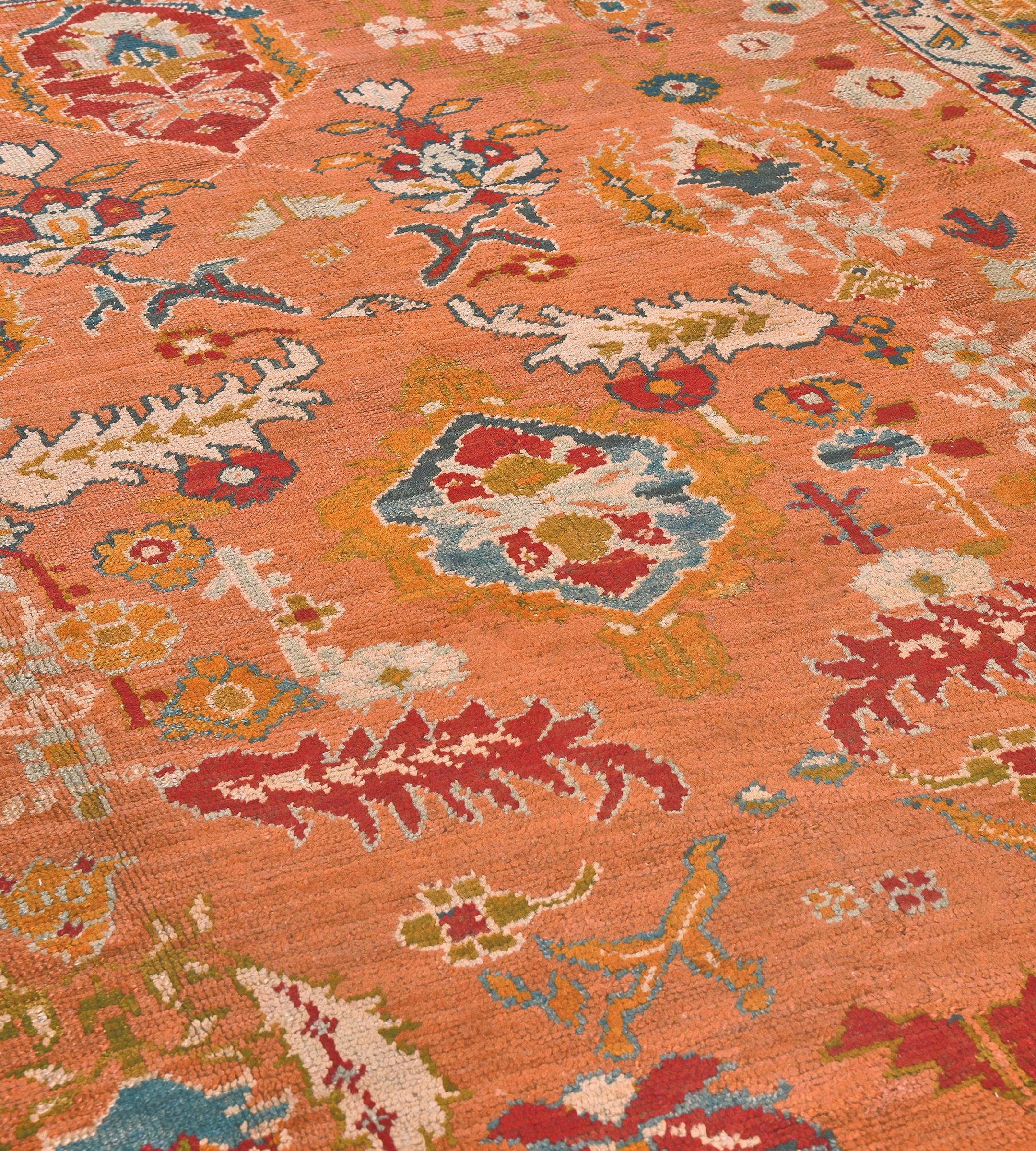 This antique Oushak Rug features a muted-orange field with an overall angular polychrome flowerheads, serrated leaves, and flowerheads, in a shaded moss-green border of angular floral vine linking bold palmettes, between soft orange, ivory and
