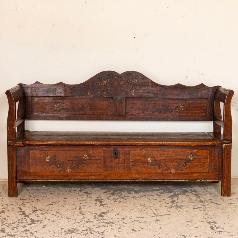 This late 19th century storage bench from Romania still maintains its original hand painted finish with a faux wood background accented with a simple flower garland motif. The hinged seat lifts to reveal ample storage inside with a divider in the