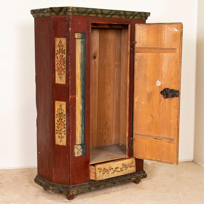 Cette charmante armoire à une porte est en excellent état d'origine. L'étui est peint en rouge brique foncé avec des bouquets floraux traditionnels peints sur le panneau avant et les côtés inclinés. Les bleus, les crèmes et les rouges présentent une