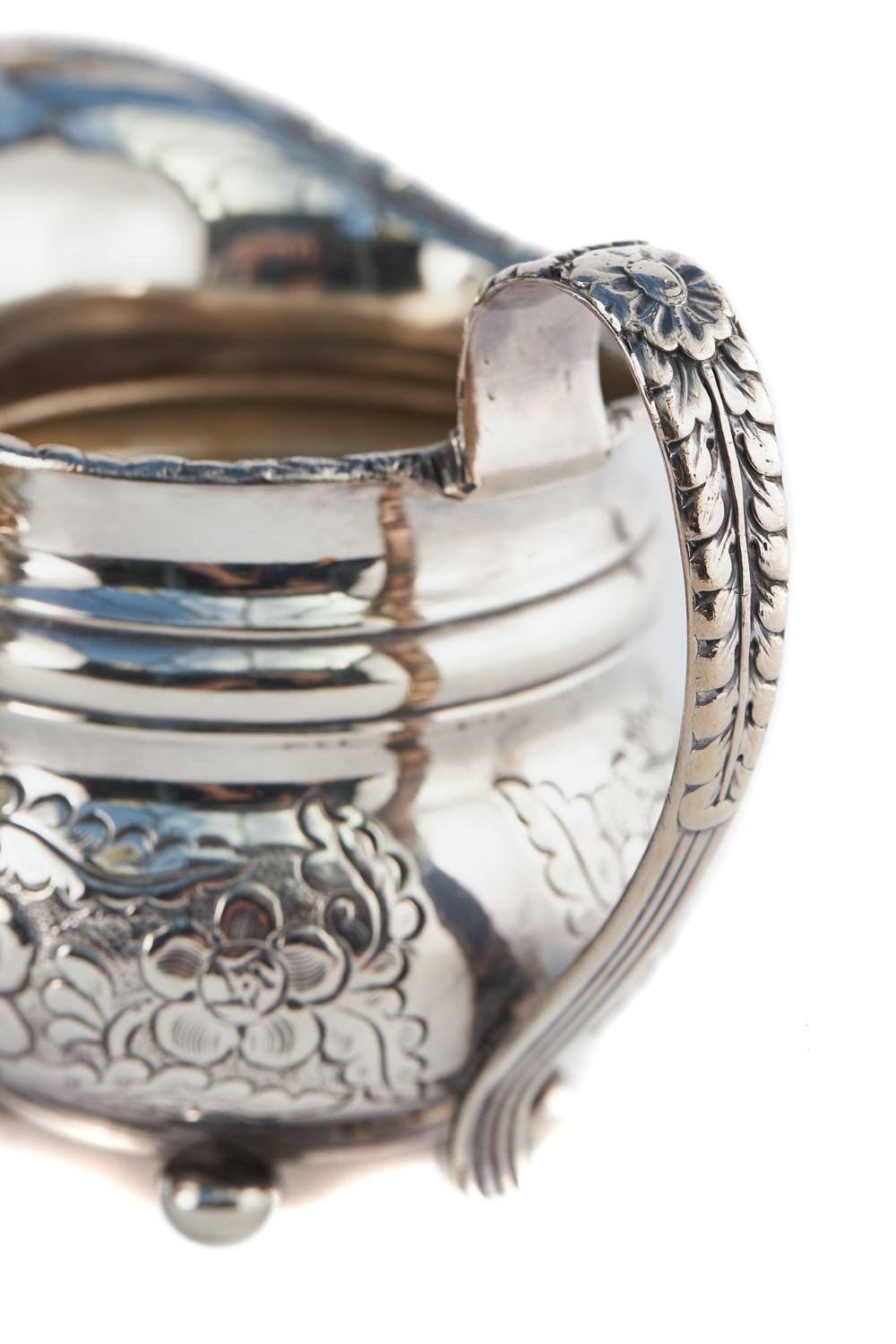 Antiguo juego de té chapado en plata ornamentada. La tetera tiene un inusual remate en forma de búho, un asa negra perfilada y se apoya en una base circular con patas de bollo. Las tres piezas del conjunto tienen la misma decoración ornamental de